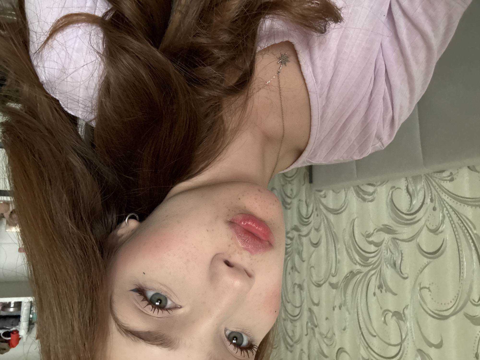 A young female is depicted from the upper chest upwards, with her head slightly tilted and her gaze directed towards the camera. She has fair skin, visible freckles across her nose and cheeks, and light blue-grey eyes accentuated with black winged eyeliner and mascara on her upper lashes. Her lips are full, pursed into a pout, and coated with a glossy pink substance. Rosy blush is applied to her cheeks. Her long, reddish-brown, wavy hair cascades over her shoulders, partially obscuring the right side of her torso. She wears a light lavender or pale purple ribbed v-neck top. A thin silver chain necklace with a star-shaped pendant hangs around her neck, and a small silver hoop earring is visible in her left ear.
The background to the left features a wall covered in light-colored wallpaper with an ornate, light-grey floral and scroll pattern. Behind her head, a light grey upholstered headboard or padded panel is visible. To the right, a white shelving unit is partially visible, containing several indistinct small objects, possibly bottles or decorative items. The lighting appears to be soft and diffuse, indicating an indoor setting.
The image does not contain any visual identifiers, such as landmarks, specific signage, or distinctive architectural elements, to confirm the location as Naberezhnye Chelny, Russia.