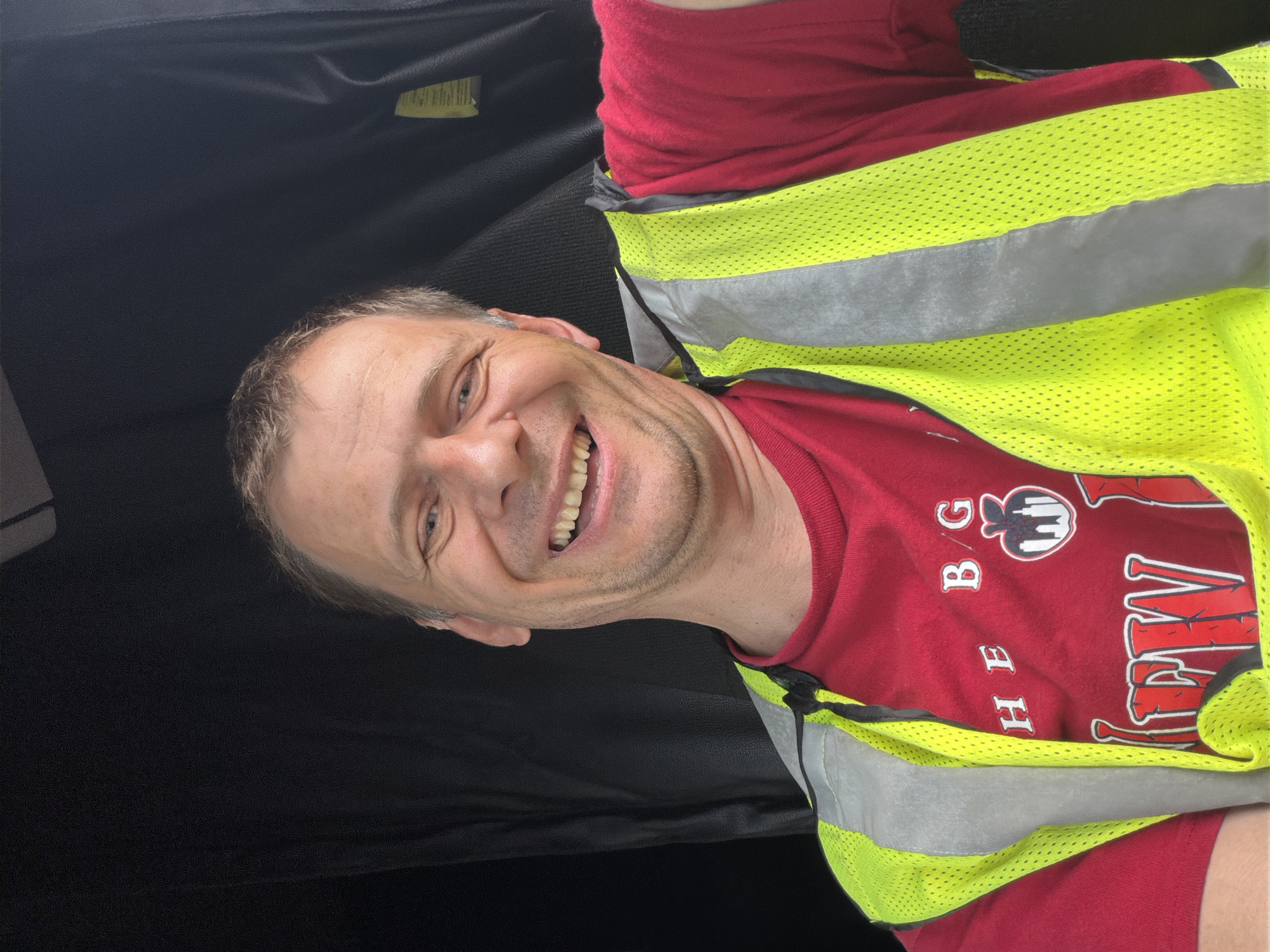 The media file is a portrait-oriented photograph of a man smiling and looking towards the camera. He is wearing a red t-shirt and a bright yellow reflective vest with gray reflective stripes. The man has short, light brown hair and stubble. He appears to be in a vehicle, indicated by the dark interior visible behind him, which is likely the headliner and a black seat or surface. The lighting suggests it is daytime. The man's expression conveys happiness or amusement. There is some text visible on his t-shirt, which appears to be a logo and some lettering. The logo is a stylized apple with a building inside, and the text reads "THE BIG APPLE." The photograph is taken at an angle, making it appear slightly tilted. The setting is likely Chattanooga, United States of America, as provided in the context. No specific weather or time of day cues are evident beyond it being daytime.