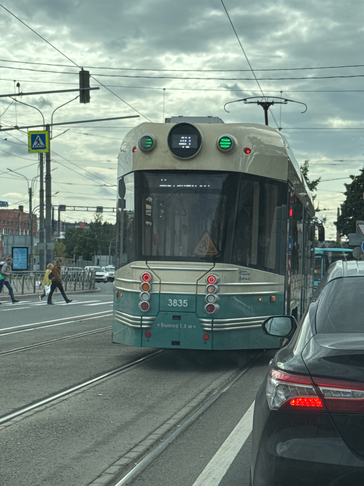 This image depicts a street scene in Saint Petersburg, Russia, on a cloudy day. The focal point is a modern, cream and teal tram, numbered 3835, with its front facing the viewer. It has distinctive round headlights that are illuminated green, and a digital display above them showing a grid of lights. The tram is positioned on tram tracks in the foreground.
To the left of the tram, a pedestrian crossing is visible, with three pedestrians in motion. One person, wearing a yellow jacket and dark pants, is walking across the street from right to left. Another person, in a brown jacket and dark pants, is also crossing from right to left, a bit behind the first. A third person, wearing a light green bag and dark pants, is walking across from left to right.
Behind the tram, a black car is partially visible, with its taillights illuminated red. In the background, there are buildings, trees, streetlights, and overhead power lines for the tram and trolleybus systems. A blue trolleybus is also visible further down the street. The overall atmosphere is urban and busy, with overcast skies suggesting a potentially damp or cool day. The text below the tram number reads "Вынос 1,5 м", which likely refers to a clearance measurement.