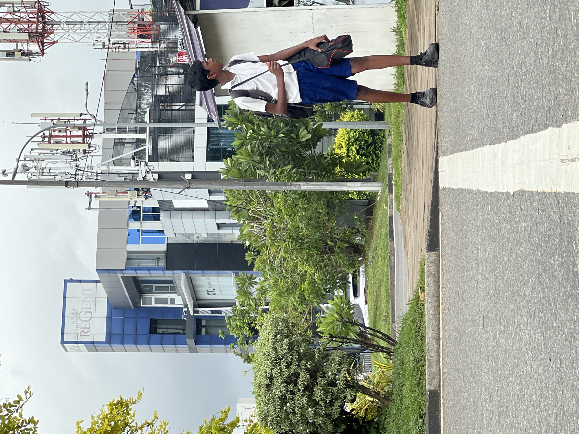 A young boy stands on a sidewalk next to a street in an urban area. He is wearing a white short-sleeved shirt, blue shorts, and black shoes. He has a black backpack slung over his left shoulder and is holding a black bag with red trim in his right hand. The boy is looking to his right. Behind him, there is a modern building with blue and gray accents, and a large sign that reads "THE REGENT". Further in the background, there are several tall telecommunication towers. Green trees and bushes are visible on the left side of the image, lining the sidewalk. The ground is paved with asphalt, and there is a white line painted across it, indicating a crosswalk. The sky is overcast.