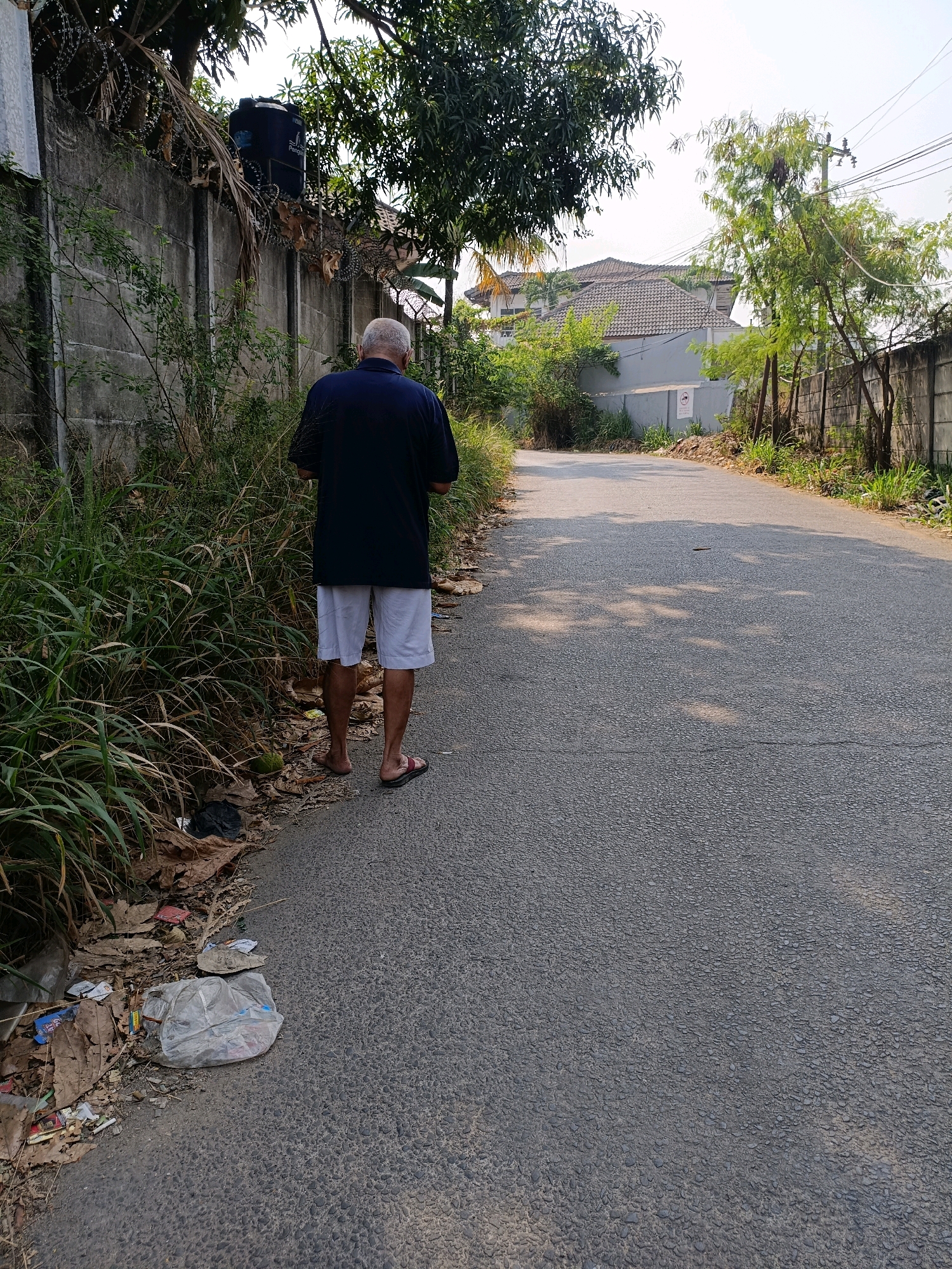 The image depicts a man standing on the side of a road in Tangerang, Indonesia. He is facing away from the camera, wearing a dark blue short-sleeved shirt and white shorts. His legs are bare, and he is wearing dark-colored sandals. The road is asphalt and appears to be a residential street, with a concrete wall on the left side of the image and a chain-link fence on the right. Lush green vegetation grows along the wall. Scattered on the ground near the wall are dry leaves and some litter, including a plastic bag. Beyond the wall and fence, buildings with tiled roofs are visible. The sky is bright, suggesting it is daytime. The overall scene conveys a mundane urban or suburban environment.