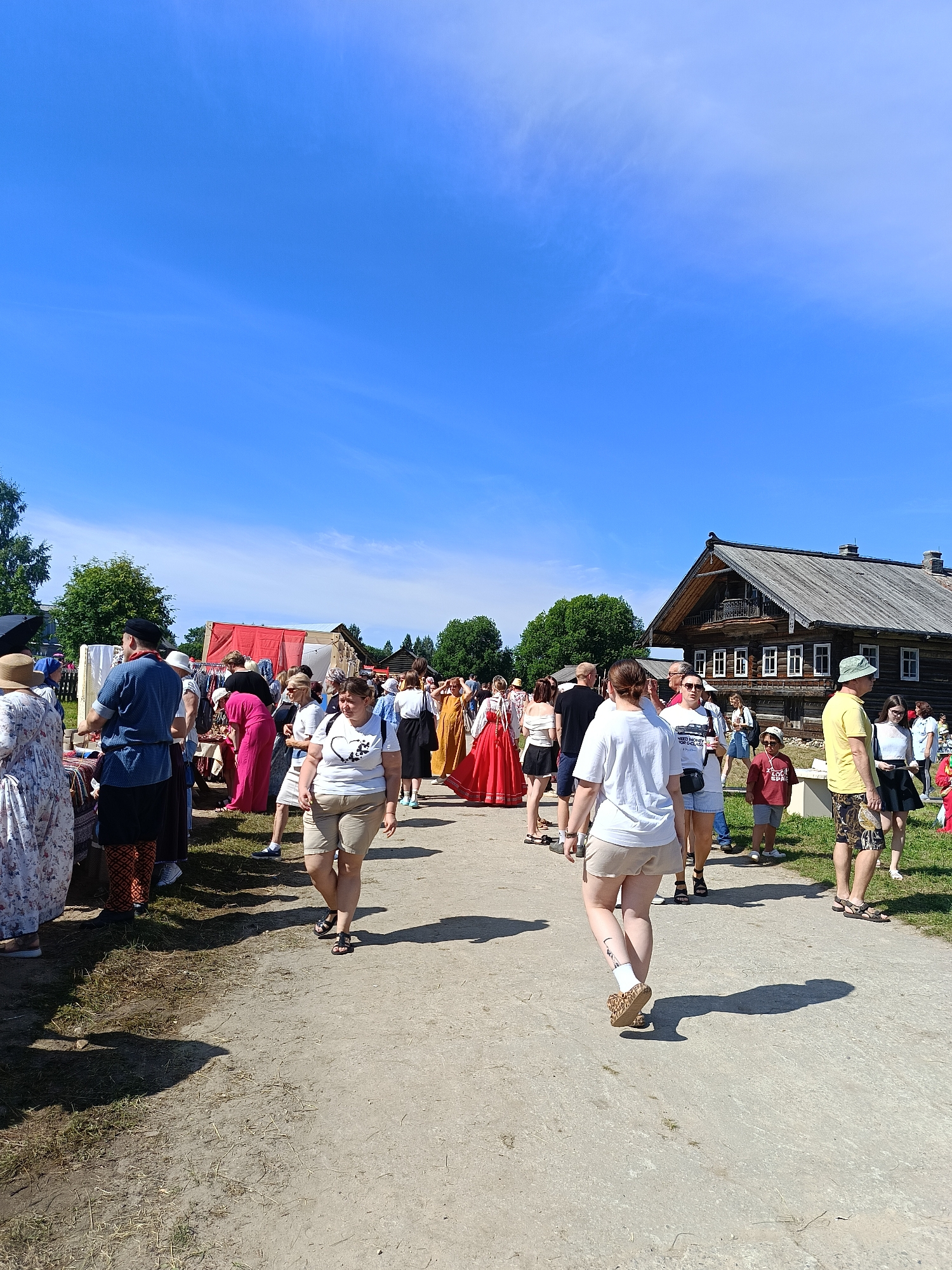 The image captures an outdoor festival or market event in Semenkovo, Russia, under a bright blue sky with scattered wispy clouds. A dirt path runs through the scene, with a crowd of people milling about. On the left side of the path, there are stalls or displays, some covered by red and white awnings, with various items laid out. People are seen browsing these displays, some dressed in modern clothing and others in what appear to be traditional or historical costumes.
Further into the scene, a large, weathered wooden building with multiple windows and a distinctive roofline, reminiscent of traditional Russian architecture, stands to the right. The building is surrounded by trees. Several individuals are walking along the path, some in groups, others individually. The general atmosphere suggests a lively, public gathering, likely a cultural or historical reenactment event given the varied attire. The ground is a mixture of dirt and sparse grass. The lighting indicates a sunny day, casting distinct shadows on the path.