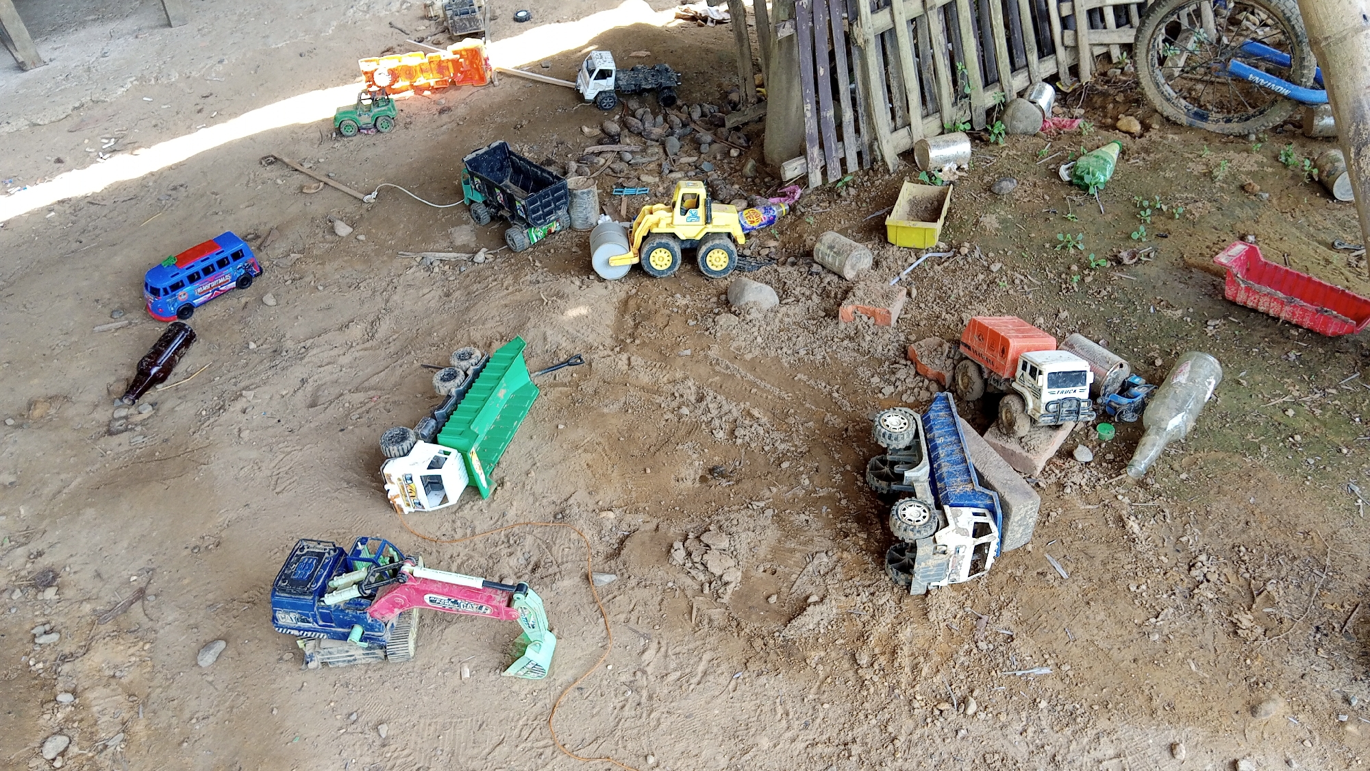 The image depicts an outdoor, somewhat disheveled scene on a dirt ground, possibly in the vicinity of Kebonbaru, Indonesia, given the context. A collection of toy vehicles, primarily trucks and construction machinery, are scattered across the ground. Among the toys are a blue and red bus, a green jeep, a black dump truck, a yellow road roller, a green flatbed truck, and a blue excavator. Several empty glass bottles, including a brown one and a larger, frosted one, are also present. A portion of a wooden fence is visible on the right side of the image, along with a bicycle wheel. The overall impression is one of a play area or discarded toys in a rustic, outdoor setting. There are no people or discernible actions captured in the frame. The environment is characterized by loose soil, small rocks, and sparse vegetation.