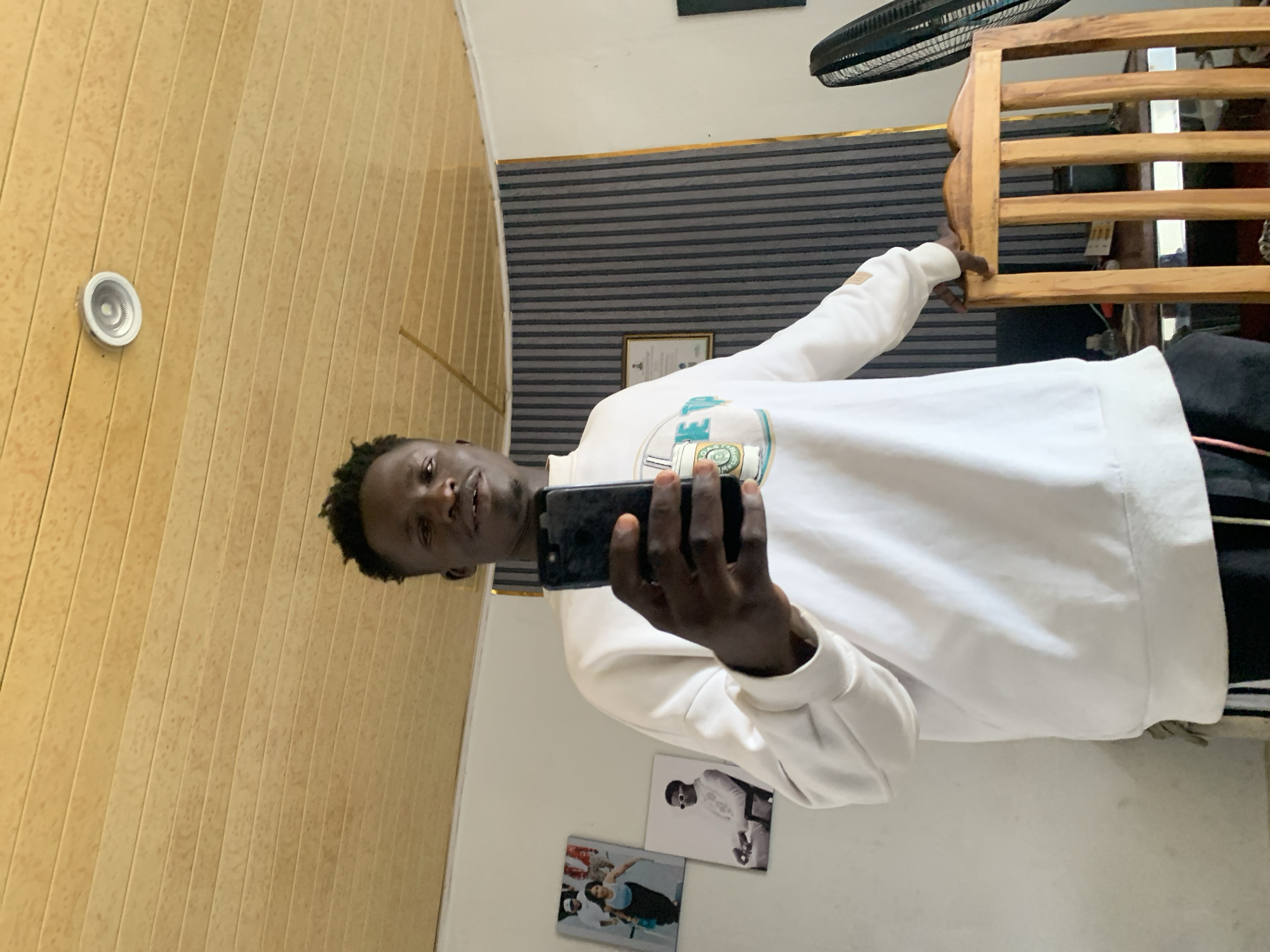 A young Black man stands in an indoor setting, holding a black smartphone in his right hand as if taking a selfie. He is wearing a white long-sleeved sweatshirt with a graphic design on the chest and black pants. He is positioned in front of a wall adorned with vertical grey and white striped wallpaper. To his left, on the wall, are two framed pictures. The ceiling above is paneled with light brown wood and features a circular light fixture. A wooden chair is visible to his right. The man's left hand rests on the back of the chair. The scene appears to be a room in a residential or casual setting within Gura Top, Nigeria.