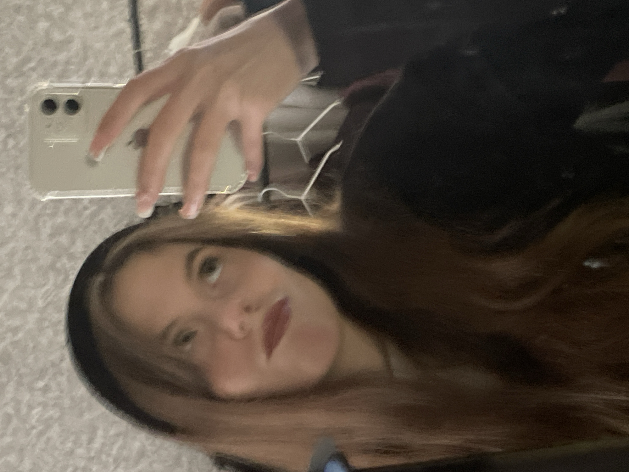A person is taking a selfie with a smartphone. The person has long brown hair, light skin, and is wearing red lipstick. The smartphone appears to be an iPhone with a clear case. The background is blurry and does not provide information about the location. The city of Фоки, Russia is not detectable from the image.