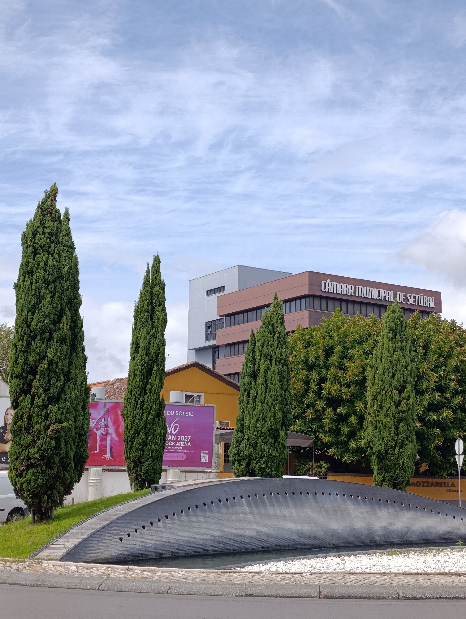 This image shows a view of the Câmara Municipal de Setúbal (Setúbal Municipal Chamber) in Setúbal, Portugal. In the foreground, there is a dark, curved structure with a series of small holes, possibly part of a fountain or a decorative element. Behind it, there are several tall, slender evergreen trees. To the left, there's a billboard advertising "CIRQUE DU SOLEIL - OVO", featuring an acrobat. Adjacent to the billboard, a white vehicle is partially visible. Further back, a yellow building with a red-tiled roof is situated. Behind these elements, the modern brown building of the Câmara Municipal de Setúbal is prominent, with its name clearly displayed in white lettering. Lush green trees surround the municipal building. The sky above is partly cloudy with a light blue hue.