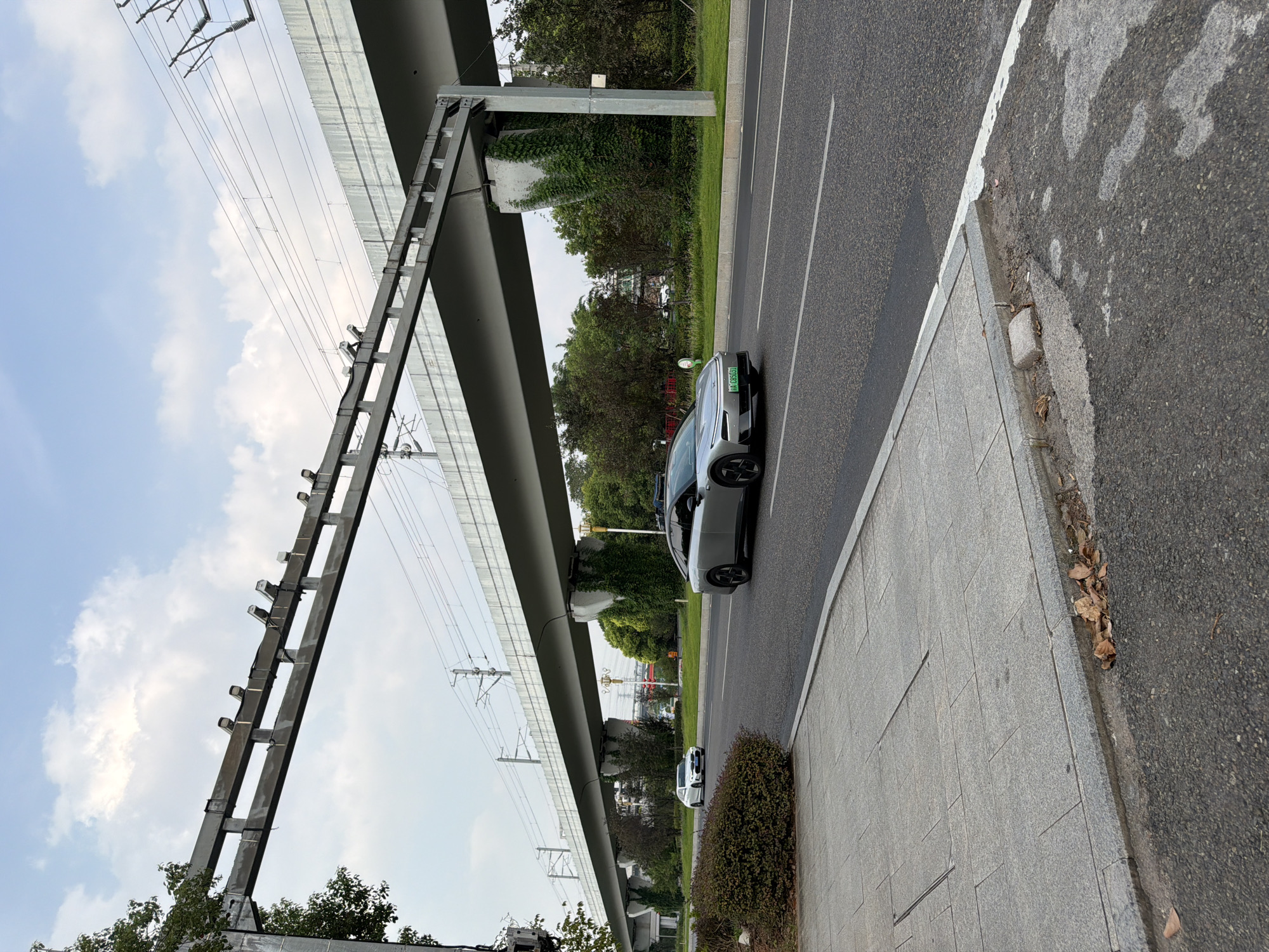 The image depicts an outdoor scene in Jiangjin, China, featuring a road with two cars. In the foreground, a modern silver sedan is driving on the right lane of the asphalt road, its front license plate reading "AA C85631". To the left and further down the road, a white sedan is also visible. Above the road, an elevated structure, likely a railway or highway overpass, spans across the scene. This overpass is supported by concrete pillars covered in green vegetation. The sky is partly cloudy with a pale blue hue. The sidewalk on the left side of the road is paved with gray square tiles, with some fallen leaves scattered on the ground near the curb. Trees and greenery are visible in the background, suggesting an urban or suburban environment.