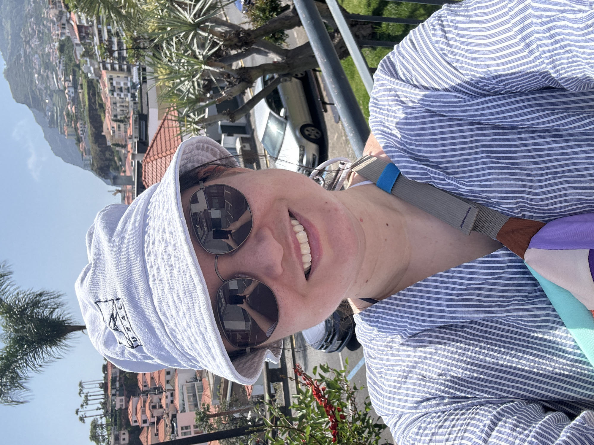A selfie shows a woman in a white bucket hat and round sunglasses, wearing a blue and white striped shirt with a grey bag strap across her shoulder. The reflection in her sunglasses shows her holding a phone to take the picture. She is smiling, revealing her teeth.
The background shows a coastal cityscape with red-roofed buildings and palm trees. A car is parked behind her, and there is a metal railing between her and the city view. Based on the distinctive coastal cityscape, red-roofed buildings and palm trees, the location is likely the city of Câmara de Lobos, Portugal. A mountain rises in the distance behind the city.