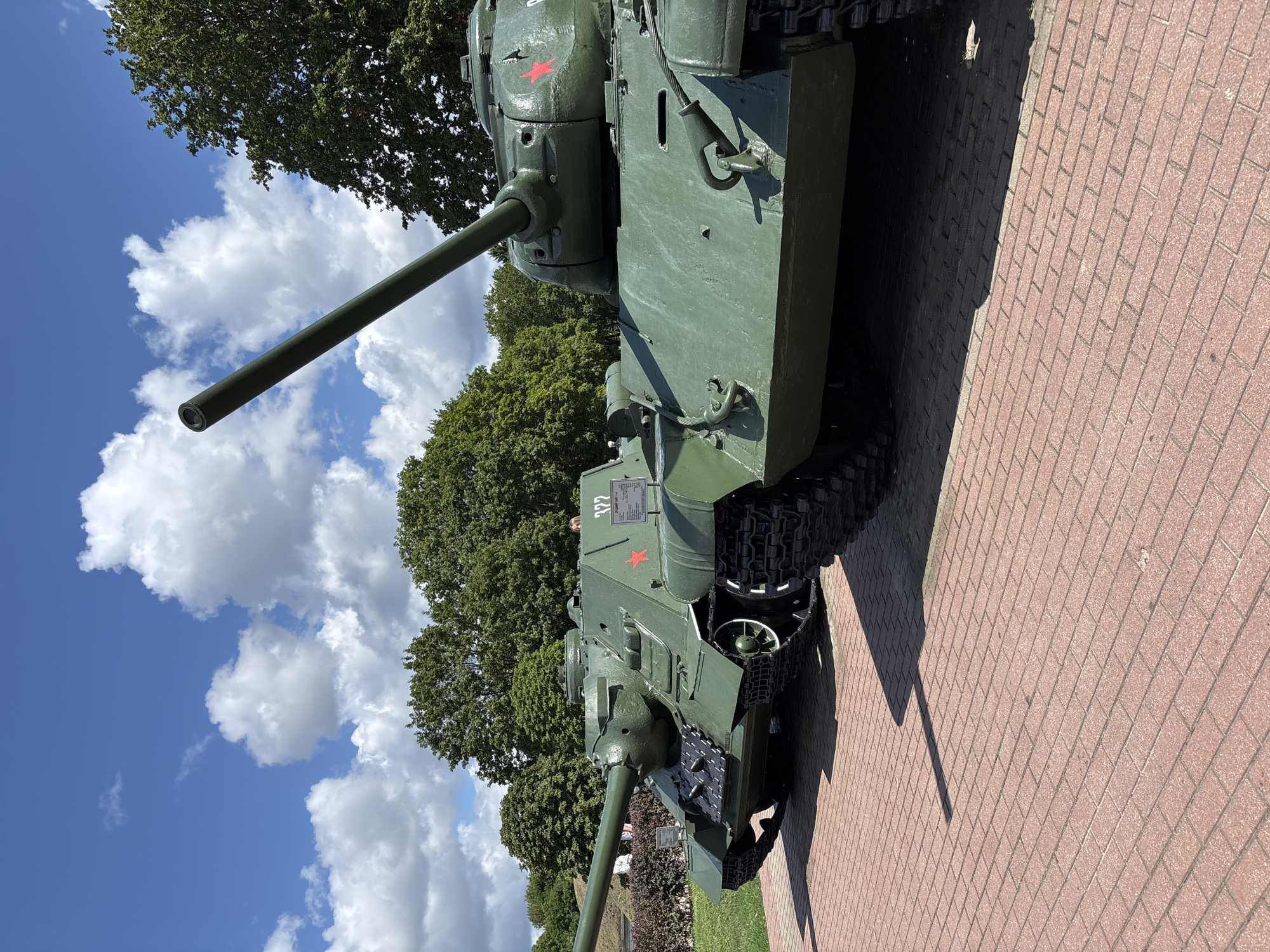 Two green Soviet-era tanks are parked side-by-side on a brick-paved area in front of lush green trees under a bright blue sky with white clouds. The tank in the foreground is closer to the viewer and is positioned at an angle, its long gun barrel pointing towards the upper right of the frame. The second tank, slightly further back and to the left, is also angled, with its gun barrel extending towards the left side of the image. Both tanks have visible tracks and turrets. There is a small information plaque visible on the tank to the left. The scene appears to be an outdoor exhibit, possibly at a memorial or museum, given the historical nature of the tanks.