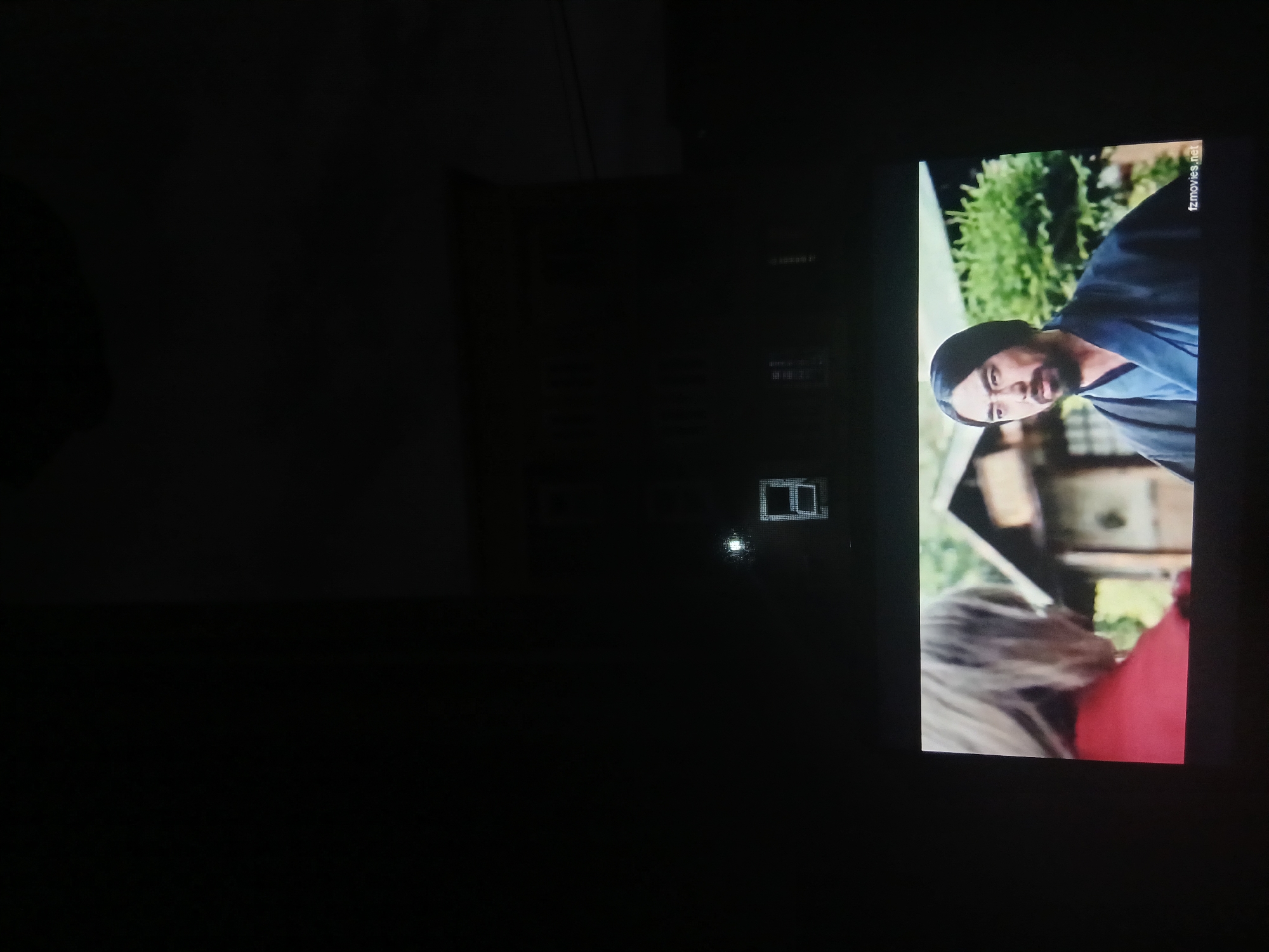 The image displays a nighttime scene from an interior vantage point in Lagos, Nigeria, observing both an exterior view and an illuminated screen. The upper two-thirds of the image are dark, revealing the silhouette of a multi-story building, likely residential, against an unlit sky. A single bright point of light, possibly from a distant source, is visible in the upper-middle left, with a distinctly illuminated rectangular window appearing below it, suggesting light either from within or reflected. The lower third of the image is occupied by a bright rectangular screen, displaying a scene from a film. On this screen, two individuals are visible: a person with light-colored hair, seen from the back or side and wearing a red garment, faces towards the right; opposite them, a man with dark hair, a beard, and a mustache, dressed in a dark, traditional kimono-style robe, looks forward and slightly left. The background of the screen content features a traditional Japanese-style wooden building and green foliage. The text "fzmovies.net" is present as a watermark in the bottom right corner of the screen. The specific street or district within Lagos is not discernible from the visual information provided.