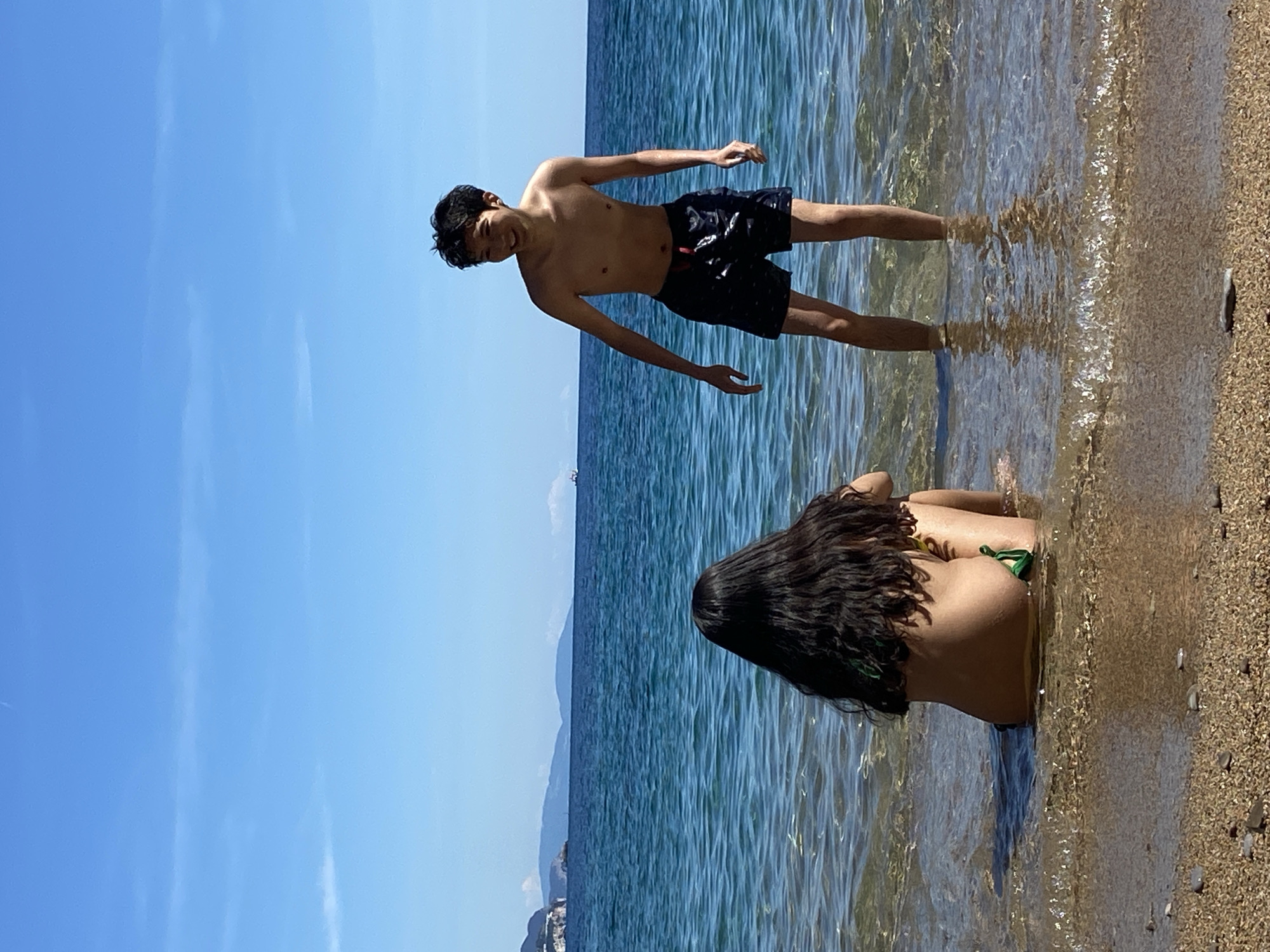 The image shows two individuals on a beach on a sunny day, with a clear blue sky. The location appears to be Budva, Montenegro. A fair-skinned male is standing in the water, wearing black shorts. The water is clear, revealing the sandy bottom. A dark-haired female is seated in the water, wearing a green bikini. The beach consists of light-colored sand. In the background, there is the sea with some distant land masses.