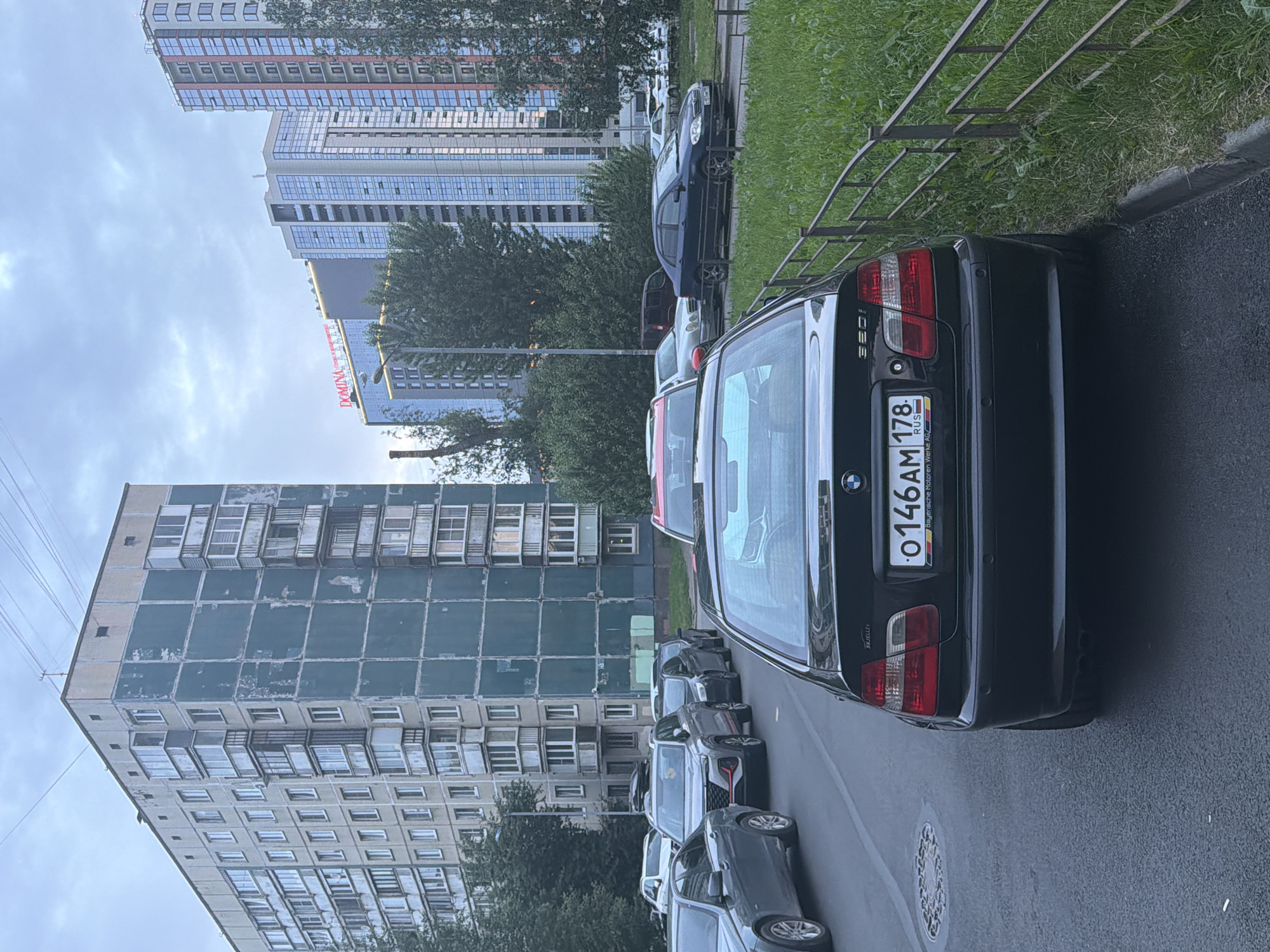 A black BMW 320i sedan is parked on a paved street in front of a grassy area bordered by a metal fence. The car has a Russian license plate that reads "O146AM 178 RUS". In the background, there are several multi-story residential buildings and modern high-rise buildings. The sky is overcast. A manhole cover is visible on the street to the left of the BMW.