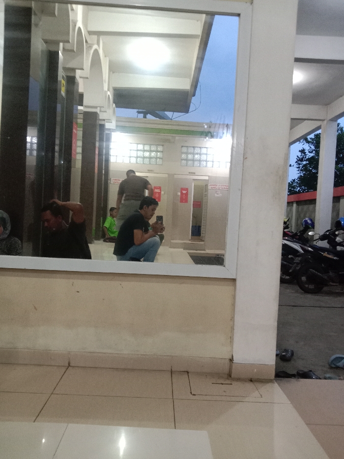 The image depicts a public or semi-public facility in Cabean, Indonesia, seen through a large reflective window or glass panel at an apparent twilight hour.

In the foreground, reflecting on the glass, a man in a black t-shirt and blue jeans is squatting, holding a black smartphone to record or photograph the scene. To his left, another man in a dark t-shirt is seated or kneeling, with his right hand touching his head. A third man in a dark t-shirt stands further back. A partial reflection of a fourth individual, possibly a woman with a head covering and checkered attire, is visible on the far left. The reflective surface is framed in white and sits atop a beige wall section, with a light-colored tiled floor extending into the immediate foreground.

Through the glass, the scene reveals an outdoor or semi-enclosed area featuring a building structure with white columns and arches, and dark vertical paneling. A bright overhead light fixture is visible on the underside of a white canopy or roof extension. Below this structure are two light-colored doors, likely for restrooms, each with a red sign. The door on the right displays partial Indonesian text, including "PERINGATAN DILARANG" (Warning: Do not) and "SAMPAH" (trash), suggesting a no-littering directive. The left door has a sign indicating "PRIA" (Men). Above the doors, glass block windows are visible. A small child in a green shirt is present in this area.

To the far right, beyond a white structural column, an outdoor area with a dark paved surface is visible. Several motorbikes are parked, and multiple pairs of sandals and shoes are scattered on the ground, indicating people have removed their footwear. The background features a red-topped wall and dark foliage against a light blue, twilight sky.