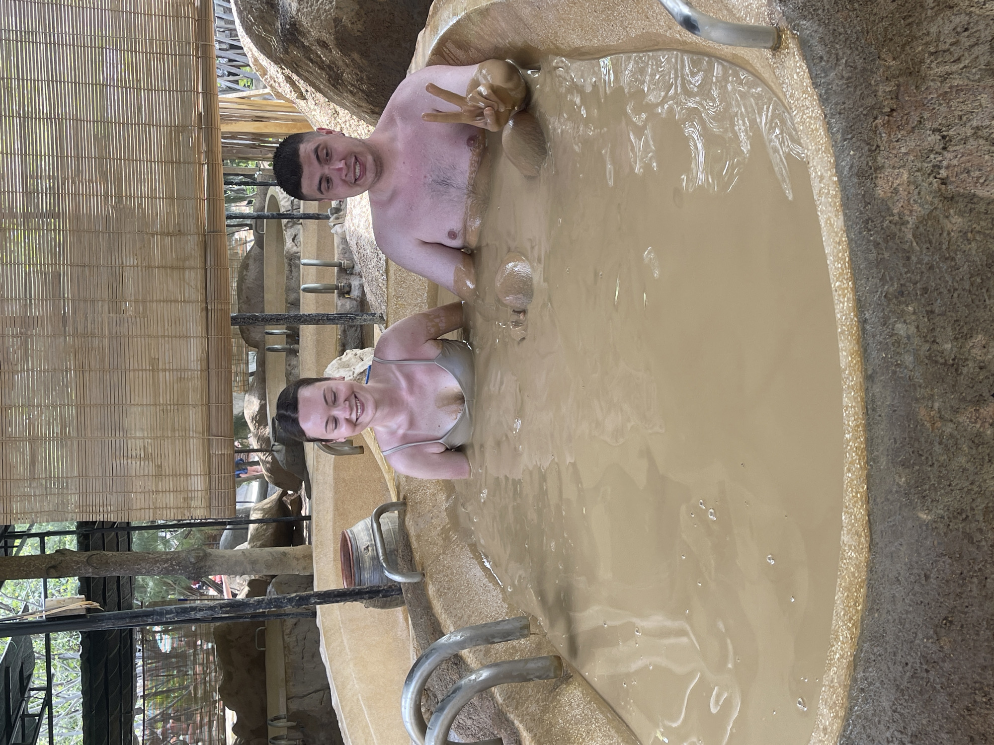 Two people, a man and a woman, are immersed in a mud bath. The man is giving the peace sign with his right hand. They are in a round stone tub filled with light brown mud. The location is outside in a natural environment with bamboo blinds and stone structures visible in the background. The photograph was taken in Tây Nha Trang, Vietnam.