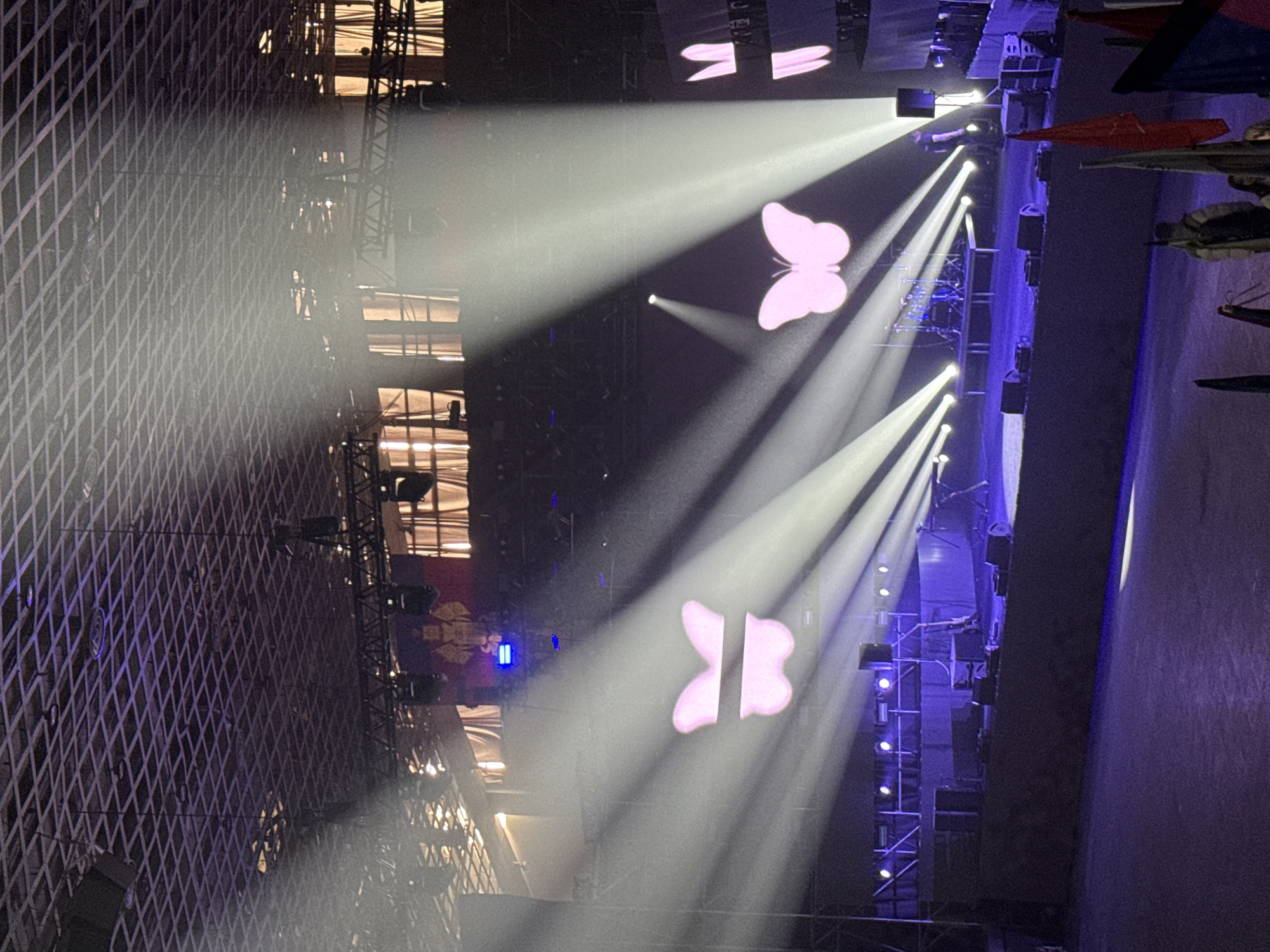 The image shows a concert stage with bright white spotlights shining through the air, forming a butterfly shape in the light. The stage is constructed of metal scaffolding and a mesh wall. Several speakers and lights are visible. The image appears to have been taken from the audience.
