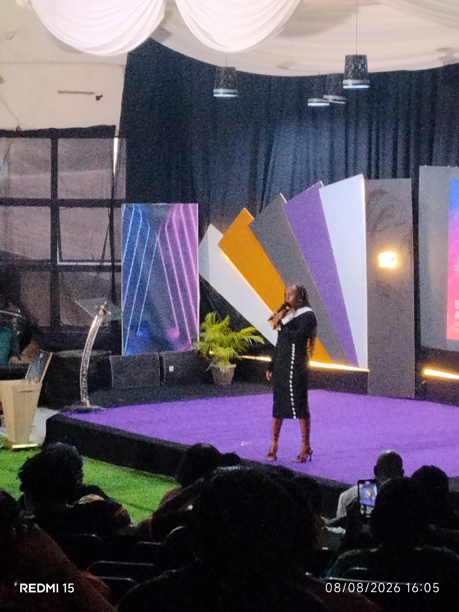 A woman in a black dress and brown heels stands on a purple stage, holding a microphone and speaking. She is facing the left side of the image, with her hair down her back. Behind her, a black curtain serves as a backdrop. To her left is a podium. To her right, a large screen displays abstract colorful shapes. The stage is illuminated by purple lighting and some yellow stage lights. In the foreground, the audience is visible, silhouetted against the stage lights. The bottom left corner of the image has the text "REDMI 15", and the bottom right corner shows "08/08/2026 16:05". The image appears to be taken in an indoor venue, possibly a conference hall or auditorium, in Aba, Nigeria.