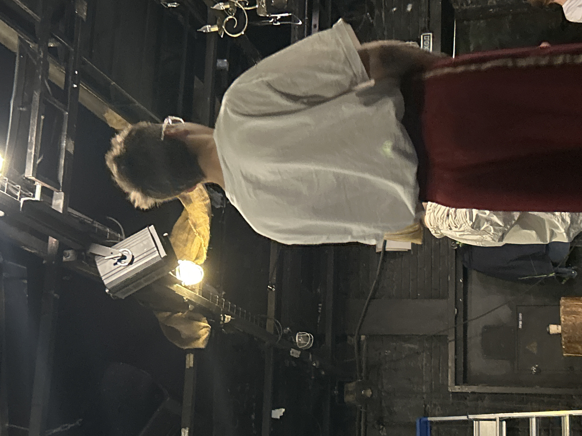The image shows the back of a person from the waist up. The person is wearing a light-colored t-shirt and dark red pants with a stripe down the side. They have short, dark, messy hair and appear to be wearing glasses. The background is dark and appears to be an indoor space, possibly a theater or backstage area, with various lighting fixtures and structural elements visible overhead. There is also a ladder and a door in the background. The overall lighting is dim.