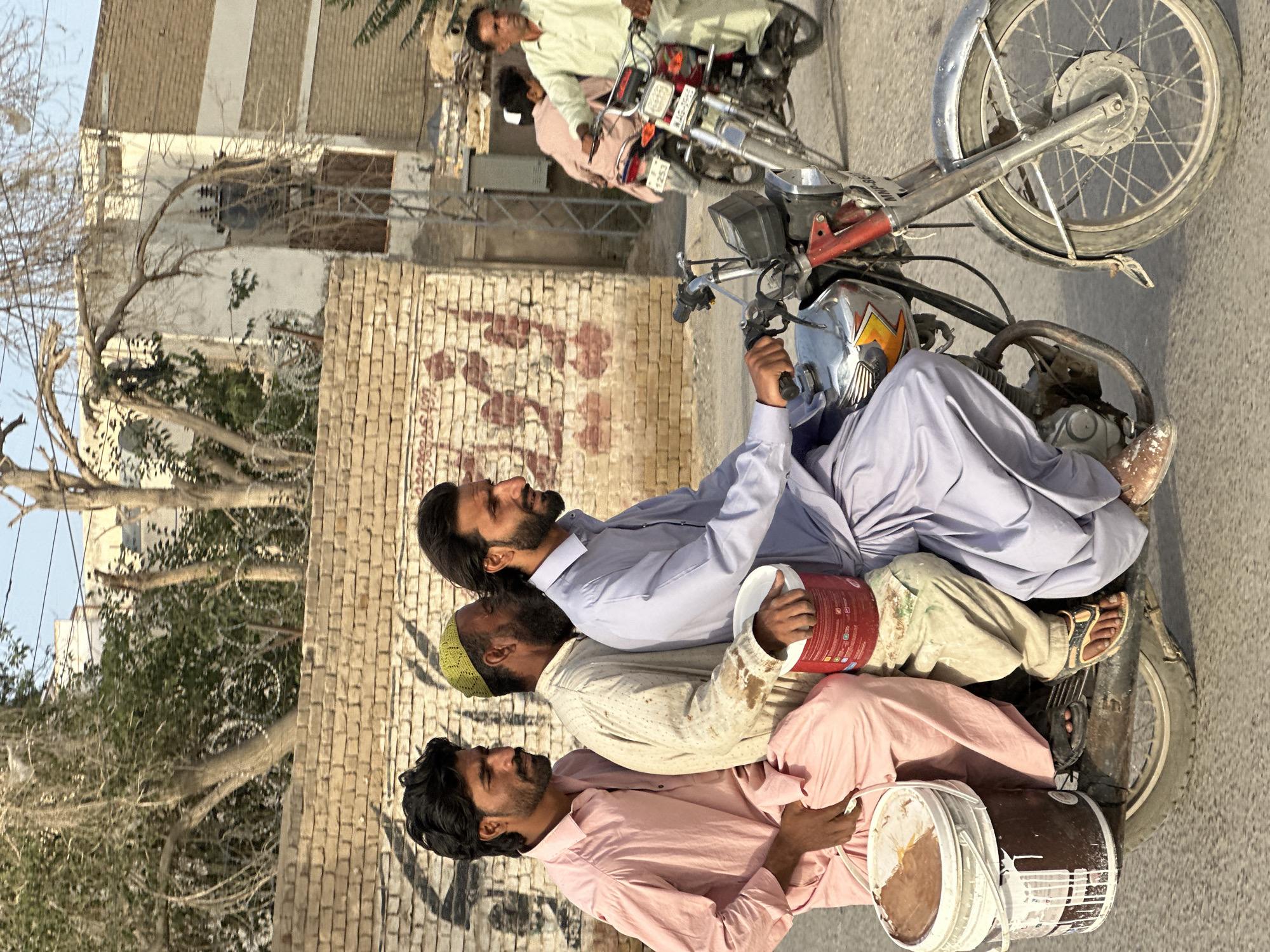Three men ride a motorbike in Quetta City Tehsil, Pakistan. The driver wears a light blue shirt, and the man in the middle wears a cream shirt and a green cap. The third man wears a pink shirt and holds a paint bucket. The second man also holds a paint bucket. Another motorbike with two people sits behind them. A brick building is in the background. Trees are on the left side.