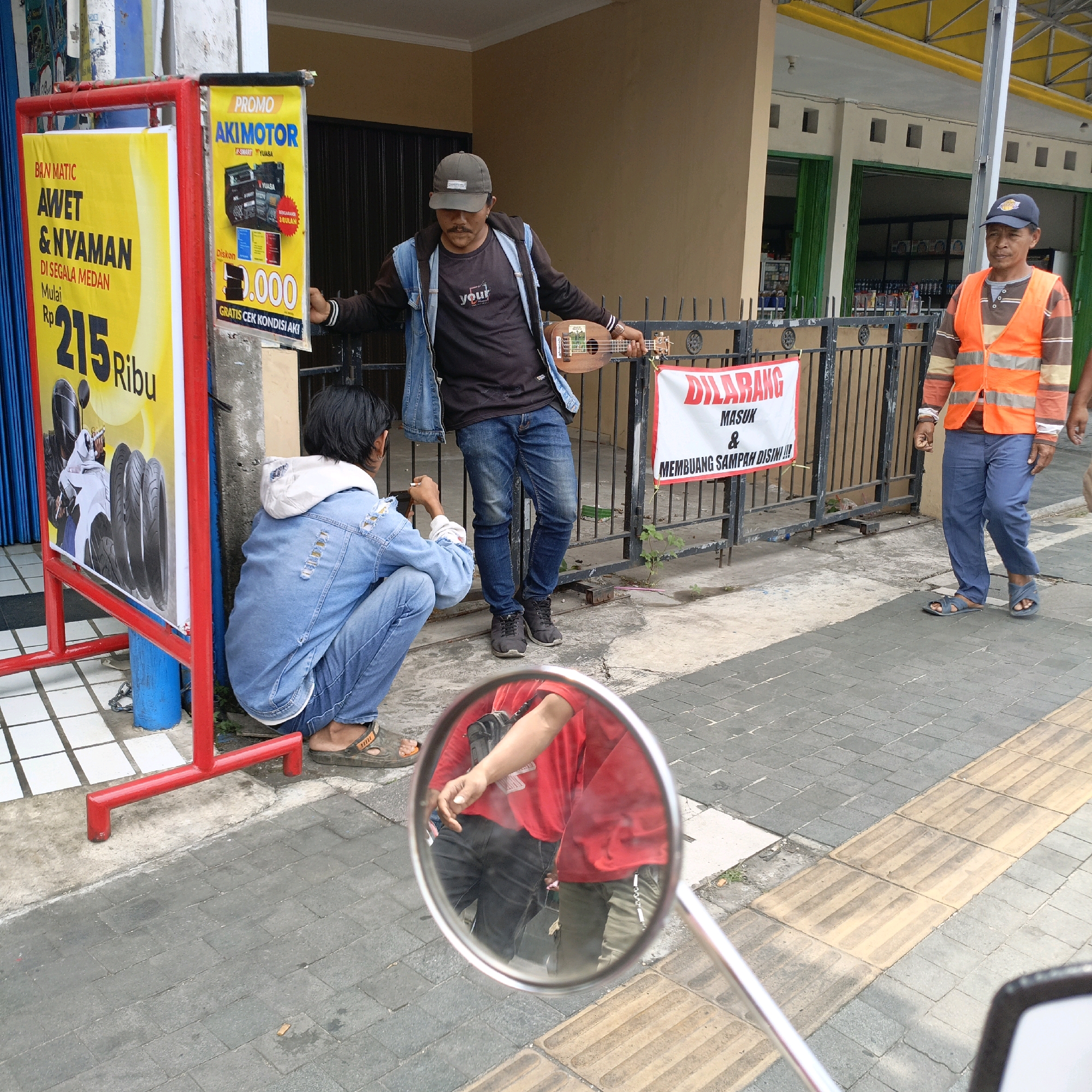A wide shot captures a street scene in Salatiga, Indonesia, focusing on several individuals near a storefront. In the foreground, a red metal A-frame sign stands on a checkered tiled surface, displaying yellow promotional posters. One poster advertises "BRN MATIC AWET & NYAMAN DI SEGALA MEDAN" with a price of "Rp 215 Ribu" and images of tires and a motorcycle. To the right, another yellow poster promotes "PROMO AKIMOTOR" featuring images of batteries.
Behind the promotional signs, a person wearing a denim jacket and jeans kneels on the sidewalk, holding a camera and appearing to take a photo. Further back, a man with a cap and mustache, also in a denim jacket and jeans, stands with a ukulele in his hand, leaning against a metal fence. A white banner with red lettering that reads "DILARANG MASUK & MEMBUANG SAMPAH DISINI!!!" is attached to the fence. To the far right, a man in an orange reflective vest and blue pants walks away from the camera, looking towards the viewer. The background shows a building with green doors and yellow trim, suggesting it is a commercial establishment. The ground is paved with grey and yellow tiles.
