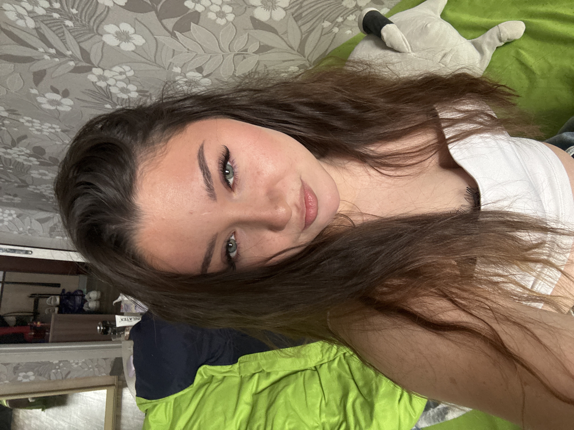 The image is a close-up selfie of a young woman with long, brown hair and green eyes, looking directly at the camera. She is wearing a white tank top. Her makeup includes eyeliner, mascara, and blush. She is leaning back on a bright green surface, possibly a bed or sofa, with a patterned wallpaper visible in the background. A stuffed animal is partially visible to her right. The scene appears to be indoors, in a residential setting. There is no visual information in the image to indicate the specific location within the city of Perm, Russia.