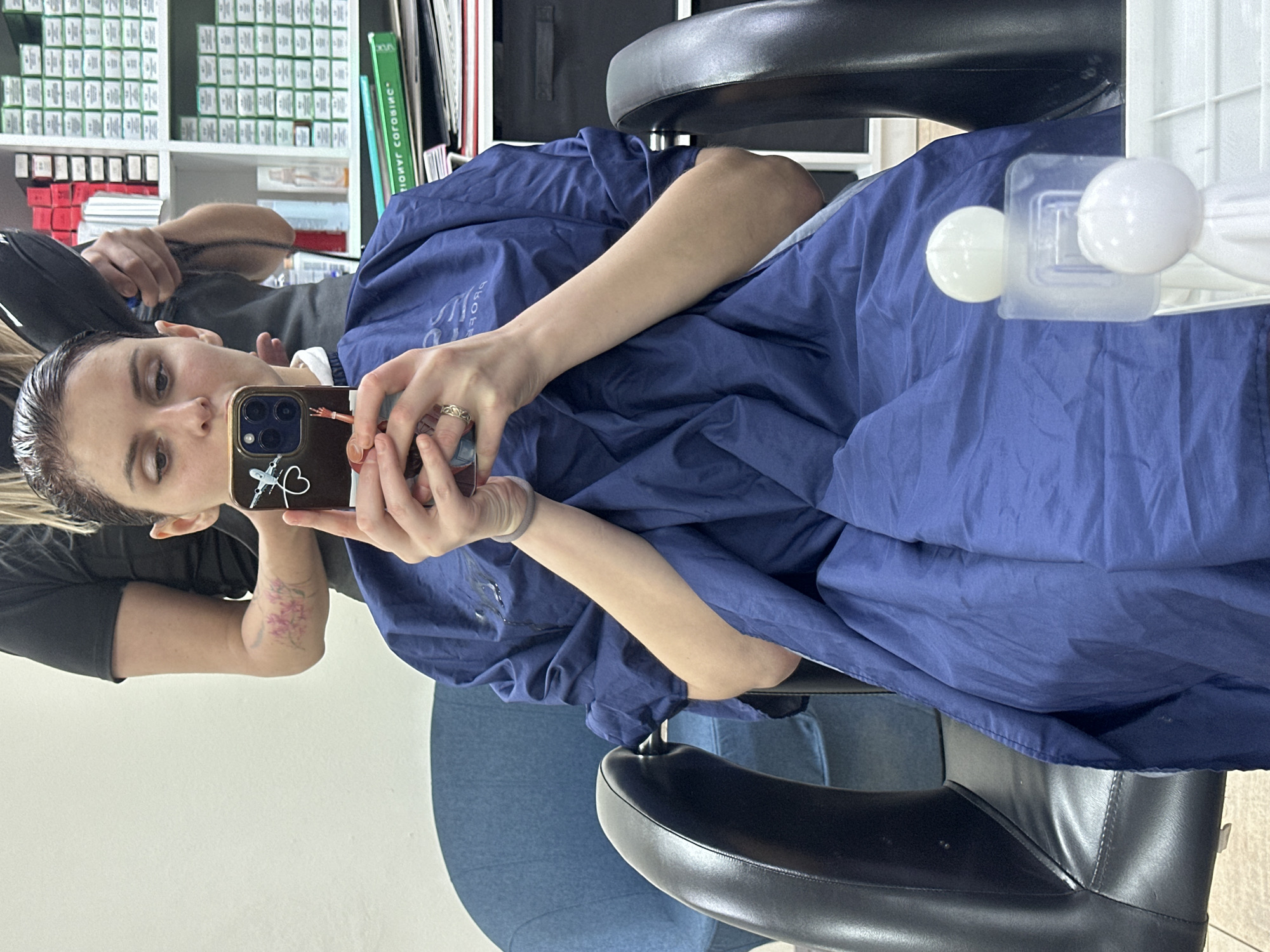 This media file is a selfie taken by a person sitting in a salon chair. The person has dark hair that appears wet and is wearing a blue cape. They are holding a smartphone to take the photo, and the phone case has a sticker of an airplane and a heart on it. The person is wearing a ring on their finger. In the background, there are shelves filled with hair products, likely hair dye boxes. There are also some green books or pamphlets on a shelf. The scene appears to be indoors, in a salon setting. The lighting suggests it could be daytime. The person's expression is neutral. There is no visible text on the person, their clothing, or the surrounding objects, except for text that is potentially on the spines of the books or product boxes, which is not clearly legible.