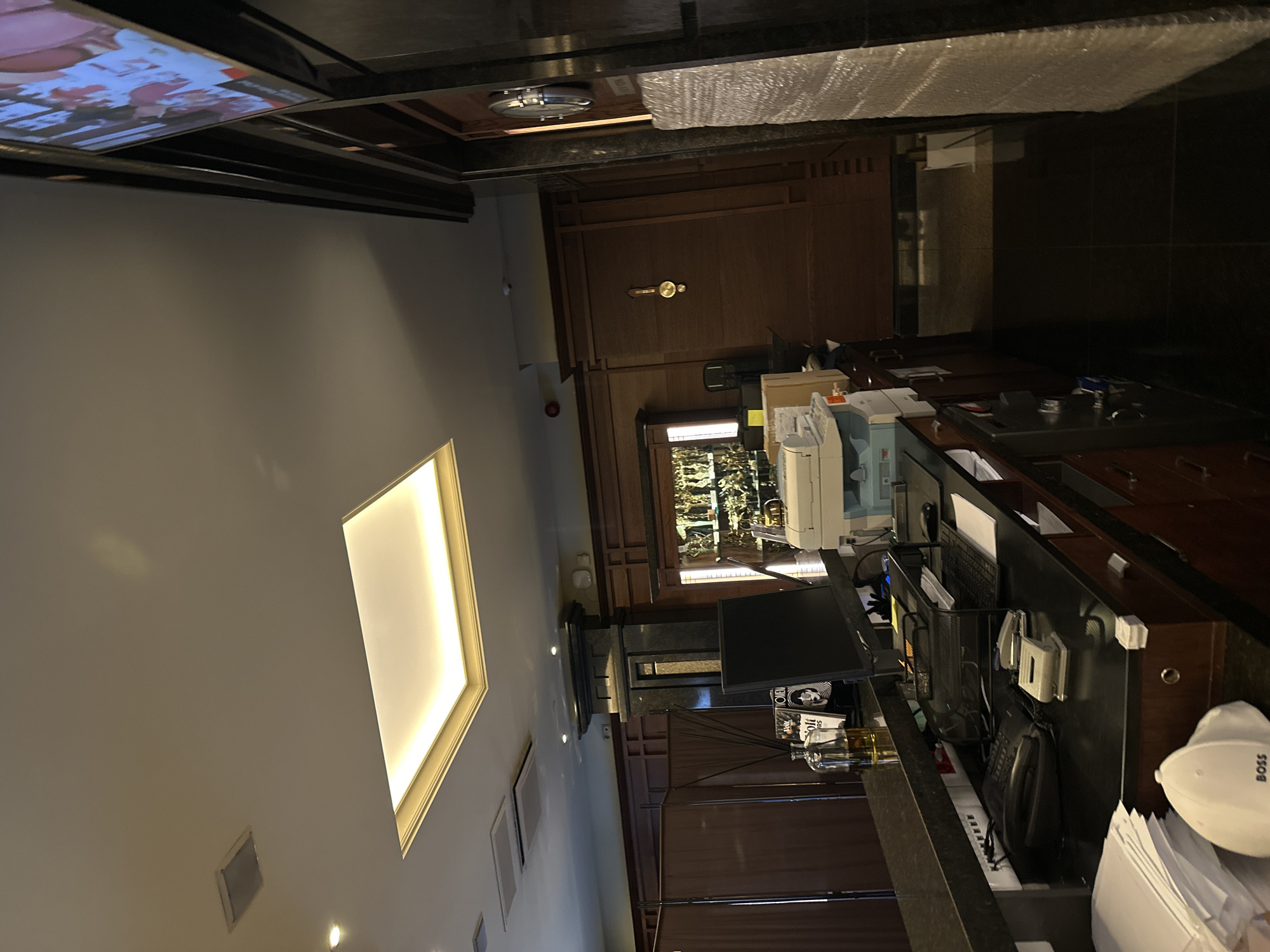The image depicts an interior scene, possibly a reception area or an office space, likely in a commercial building. The room features a high ceiling with inset lighting. A television is visible on the ceiling, showing a video. The walls are painted in a light color.
The foreground is dominated by a reception desk. The desk has a computer monitor, a keyboard, a telephone, and various office supplies. A "BOSS" branded hardhat rests on top of some papers. Behind the desk, there is a printer and filing cabinets with dark wooden veneer. A lighted aquarium serves as the backdrop behind the desk.
The room's color palette consists of neutral tones, with accents of wood and metal. A wooden door is visible in the background.
Without any additional information from the image, a definitive location in Sofia, Bulgaria is impossible to ascertain.