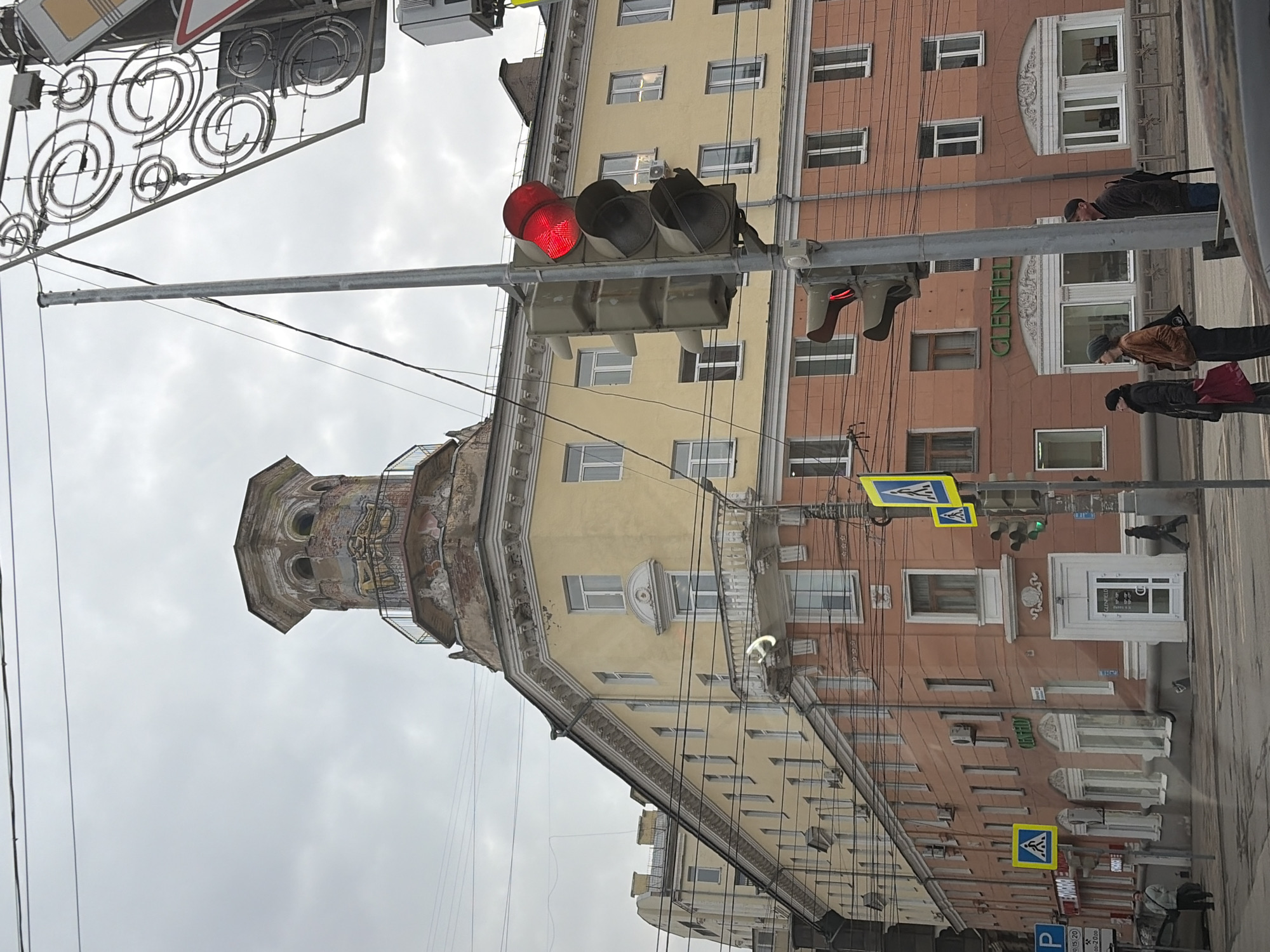 The image shows a street scene in Izhevsk, Russia. A traffic light is displaying a red light. The building adjacent to the traffic light is a multi-story structure with a mix of yellow and reddish-brown facades. Pedestrians are walking on the sidewalk next to the building. A tower-like structure with graffiti and deteriorating facade is visible in the background. Street signs are present, one of which is a pedestrian crossing sign. Overhead wires and decorative street lights are visible. The sky is overcast. The building on the right is identified as "Glenfield".