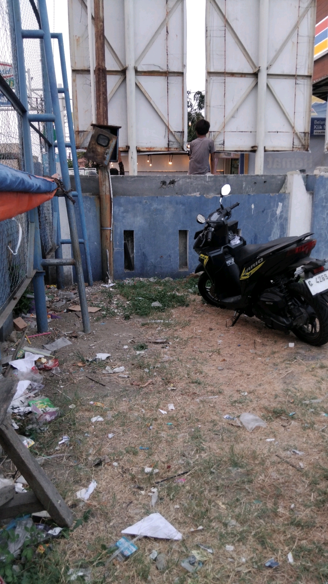 The image depicts an outdoor scene, likely in Tegal, Indonesia, characterized by a cluttered and somewhat neglected environment. In the foreground, dry grass and soil are strewn with various pieces of litter, including plastic bags, wrappers, and paper. A broken wooden plank rests at an angle in the lower left corner.
Dominating the mid-ground is a black motorcycle parked on the ground. It appears to be a scooter-style vehicle with the model name "VARIO" visible on its side. The motorcycle has a license plate that reads "G 4269". Behind the motorcycle, a blue painted wall with two rectangular openings is visible.
To the left of the motorcycle, a blue metal structure, possibly part of a fence or a sports facility, is present. Adjacent to this structure, a rusty metal pole supports a small, dilapidated electrical box.
In the background, a large, white billboard structure with metal framing is prominent. Behind this billboard, a glimpse of trees and a bright sky can be seen. A young person, seen from the back, is seated on a ledge or wall in front of the billboard, looking away from the camera. The overall impression is that of an urban fringe area, potentially an alley or a less-maintained public space.
