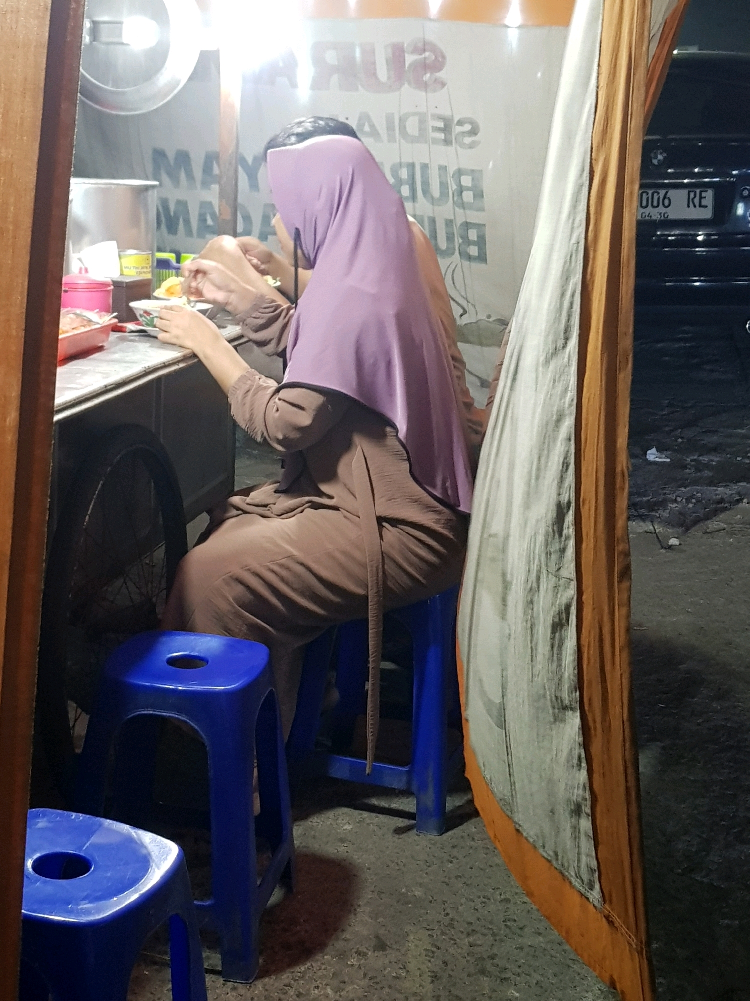 A person wearing a purple hijab and a brown garment is seated at a food stall in Bekasi, Indonesia. The person is holding utensils and appears to be eating. A bicycle wheel is visible behind the stall. The ground is paved with asphalt. A dark-colored car with a license plate reading "006 RE" is parked nearby. The scene is illuminated by artificial lighting.