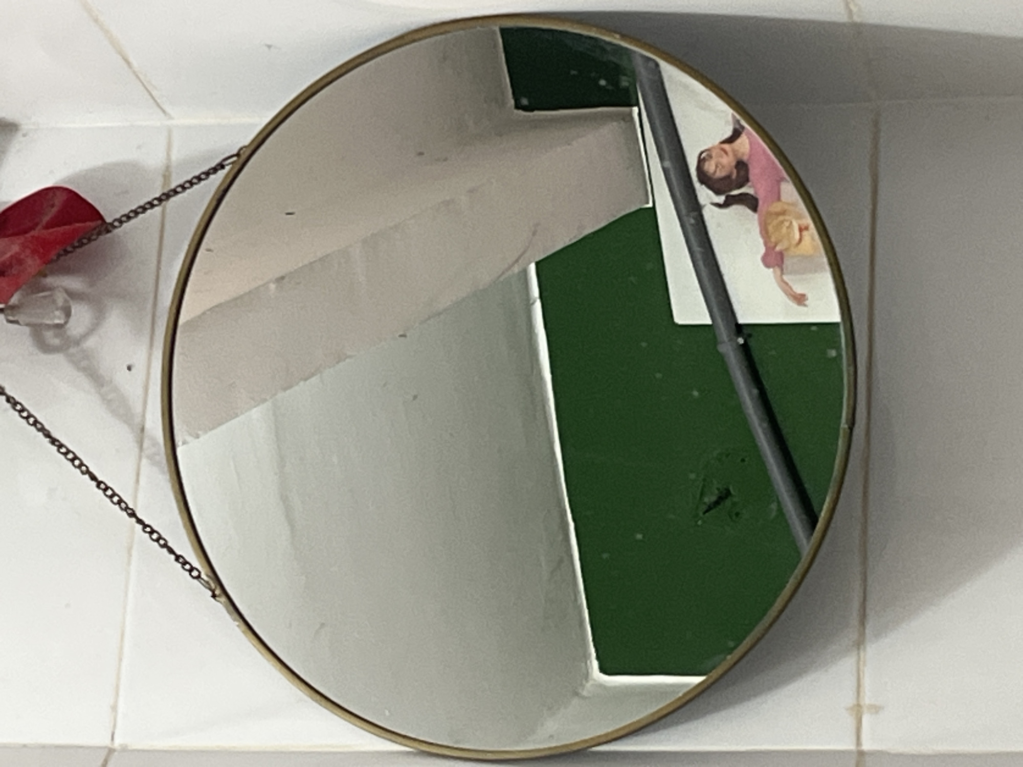 A round mirror with a thin brass frame hangs from a chain attached to the wall. The mirror reflects a corner of a room. In the reflection, a section of a green wall is visible, as is a white ceiling with a light fixture and a red decorative item hanging from it. A horizontal dark pipe runs across the green wall in the reflection. Below the pipe, a portion of a white surface is visible, on which a doll or figurine of a person wearing a pink shirt and dark pants is lying down, holding a light-colored object, possibly a toy animal, to their chest. The background wall of the room is tiled with white square tiles.