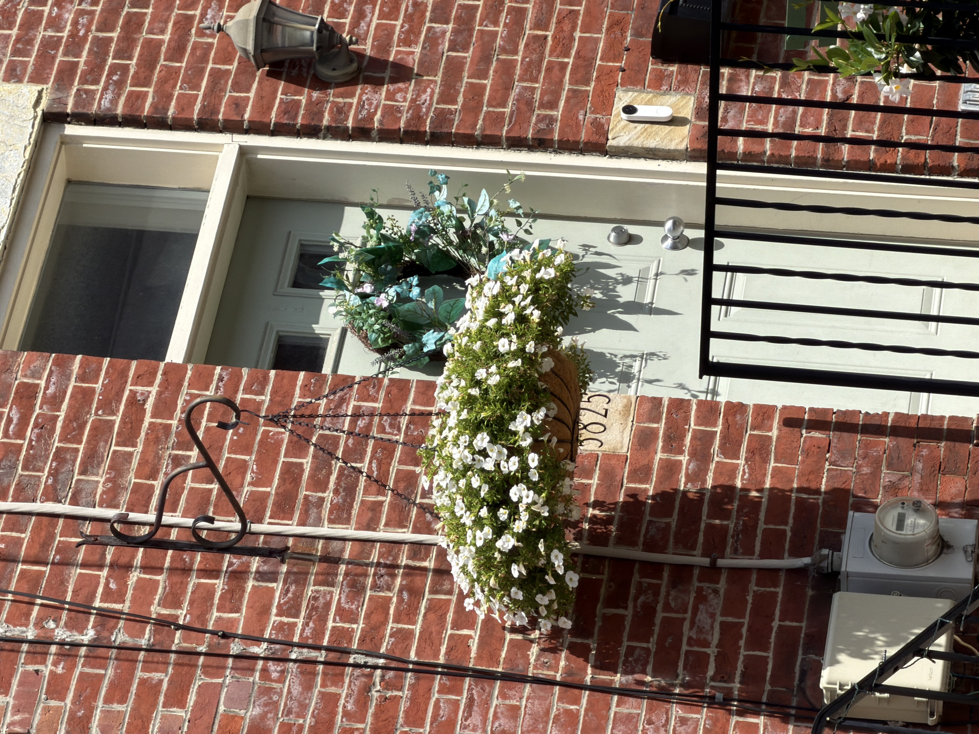 The image shows a close-up of a red brick building's exterior. A light green door is visible on the right side of the frame, with a white frame around it. Above the door, there is a window. A black metal lamppost is mounted on the brick wall to the right of the door. In front of the door, a hanging basket filled with white flowers is suspended by chains from a decorative black metal hook attached to the wall. Below the hanging basket, the house number "3825" is visible on a stone plaque. A small white doorbell camera is mounted on the wall to the right of the door. A black metal railing runs across the bottom of the image. To the left of the door, an electrical meter and junction box are attached to the brick wall. The lighting suggests it is daytime, with shadows cast by sunlight. The scene is an exterior view of a residential or commercial building in Pittsburgh, United States of America.