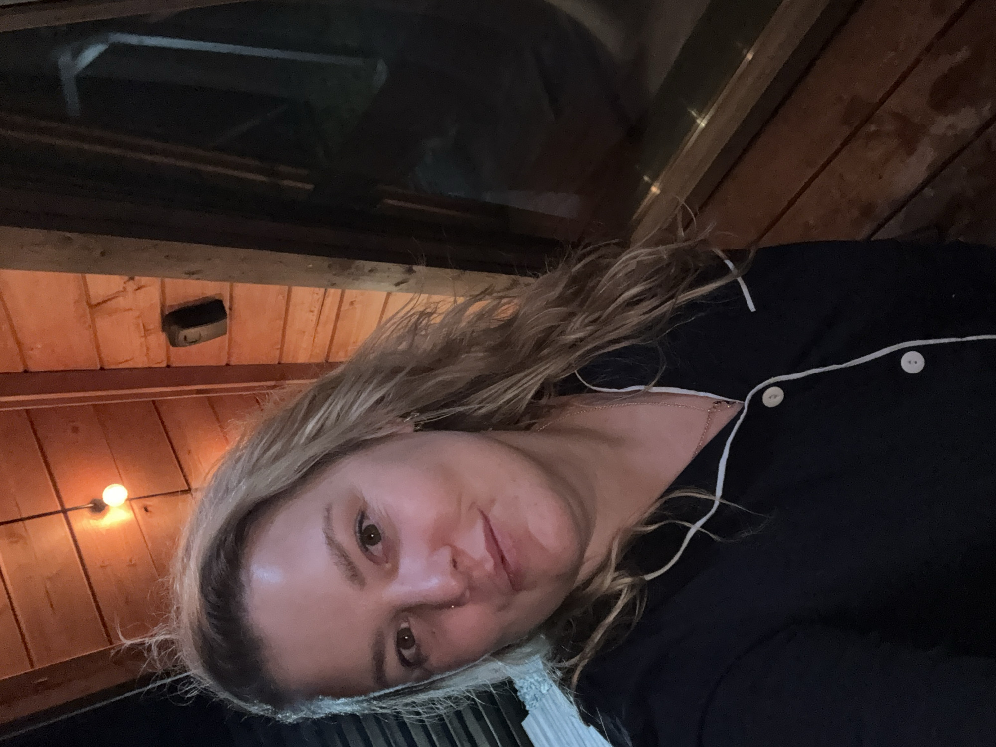 A woman with blonde hair, wearing a dark blue pajama top with white trim, is taking a selfie outdoors. She is positioned in the foreground, facing the camera with a slight smile. Behind her, a wooden wall with horizontal planks is visible, illuminated by warm lighting from an overhead bulb. To her right, a large window reflects a dark, indistinct interior space, possibly at night. The overall setting appears to be a rustic or natural environment, perhaps a cabin or porch, located in the city of Chaikovsky, Russia.