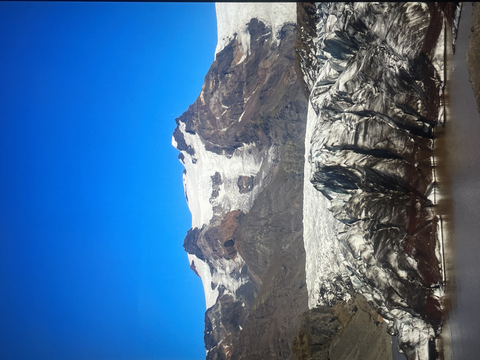 A landscape photograph showing a snow-covered mountain range on the left side of the frame under a clear blue sky. The mountain's surface is a mix of rock, ice, and patches of snow. To the right, a glacier descends from the mountain. The glacier's surface is marked with deep crevices and patterns, suggesting the movement of ice. The image is oriented vertically. The location is Unknown, Unknown.