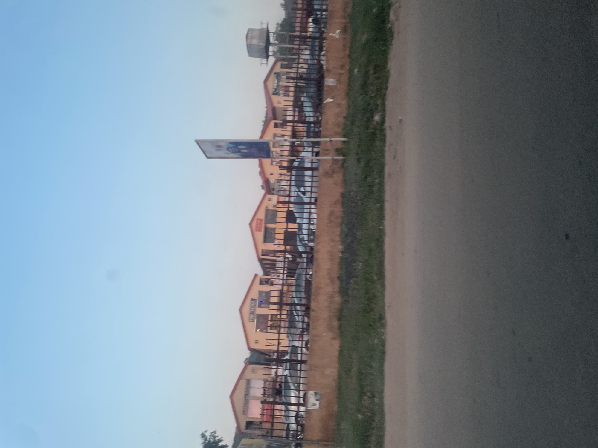 The image depicts a street scene in Abuja, Nigeria. The perspective is from the road, looking towards a row of commercial buildings. These are uniform, single-story structures with gable roofs, likely shops or businesses. A fence runs along the front of the buildings, separating them from a narrow strip of vegetation bordering the road. A tall advertising billboard is visible. A large water tank is placed above the buildings. The sky is clear and light blue, indicating either early morning or late afternoon. Several cars are parked in front of the buildings. The road is gray and takes up a substantial portion of the foreground.