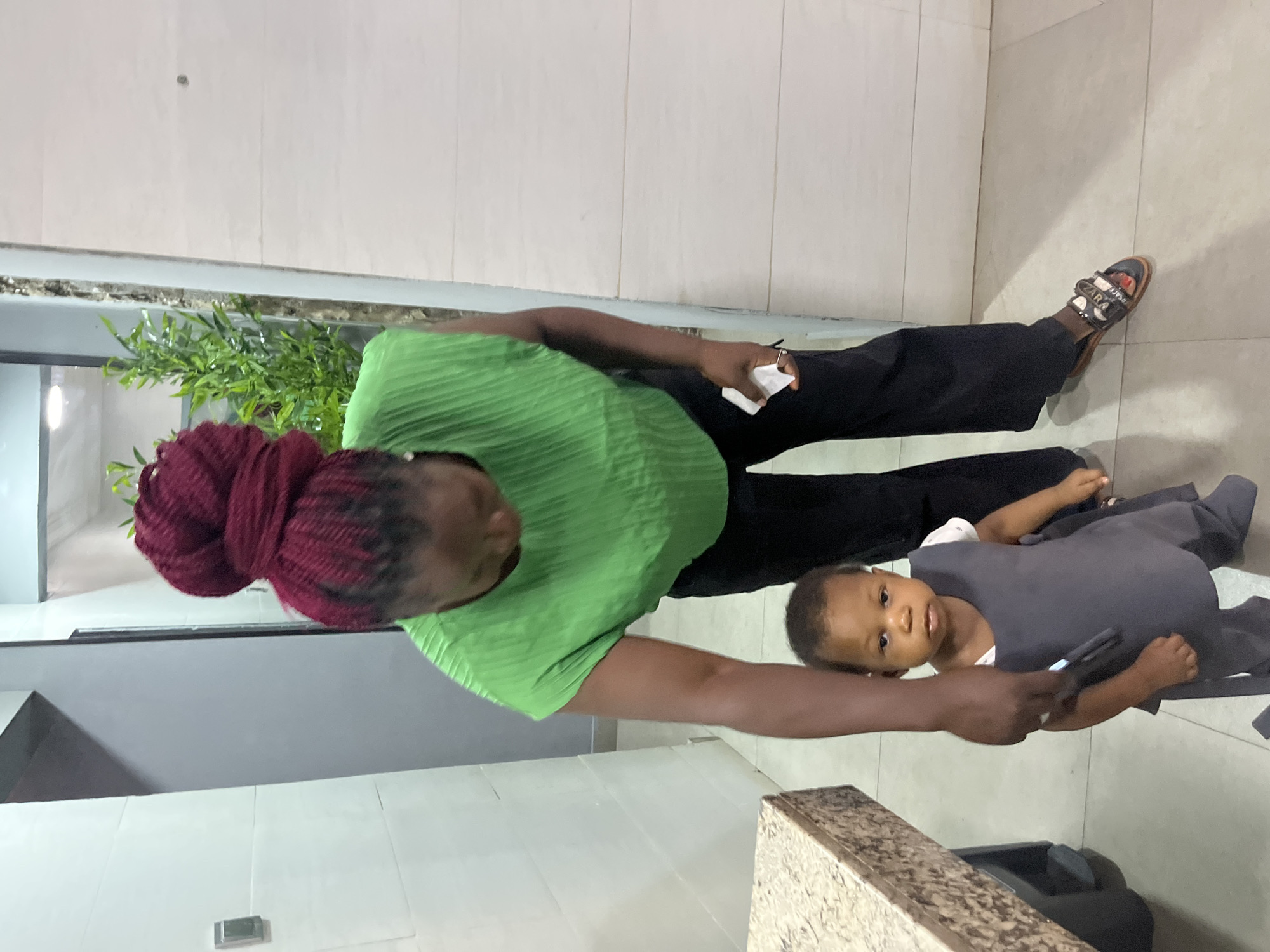 A woman with dark skin, her hair styled in a high, voluminous bun of braided, dark red extensions, wears a bright green, pleated sleeveless top and black wide-leg pants. She is bending over, her right hand extended down, holding a white object and a phone. She appears to be interacting with a young child who is standing in front of her. The child, also with dark skin and wearing a grey garment over a white undershirt, looks directly at the camera with wide eyes. The scene takes place indoors, likely in a residential or commercial space. The floor is tiled in a neutral, beige tone. To the left, a countertop with a speckled, brown and beige pattern is visible. In the background, a wall is painted a light color, and to the right, a textured, light-colored wall or paneling is present. A tall, artificial green plant stands in the background on the right. The environment suggests a modern interior. The image was taken in Abuja, Nigeria.