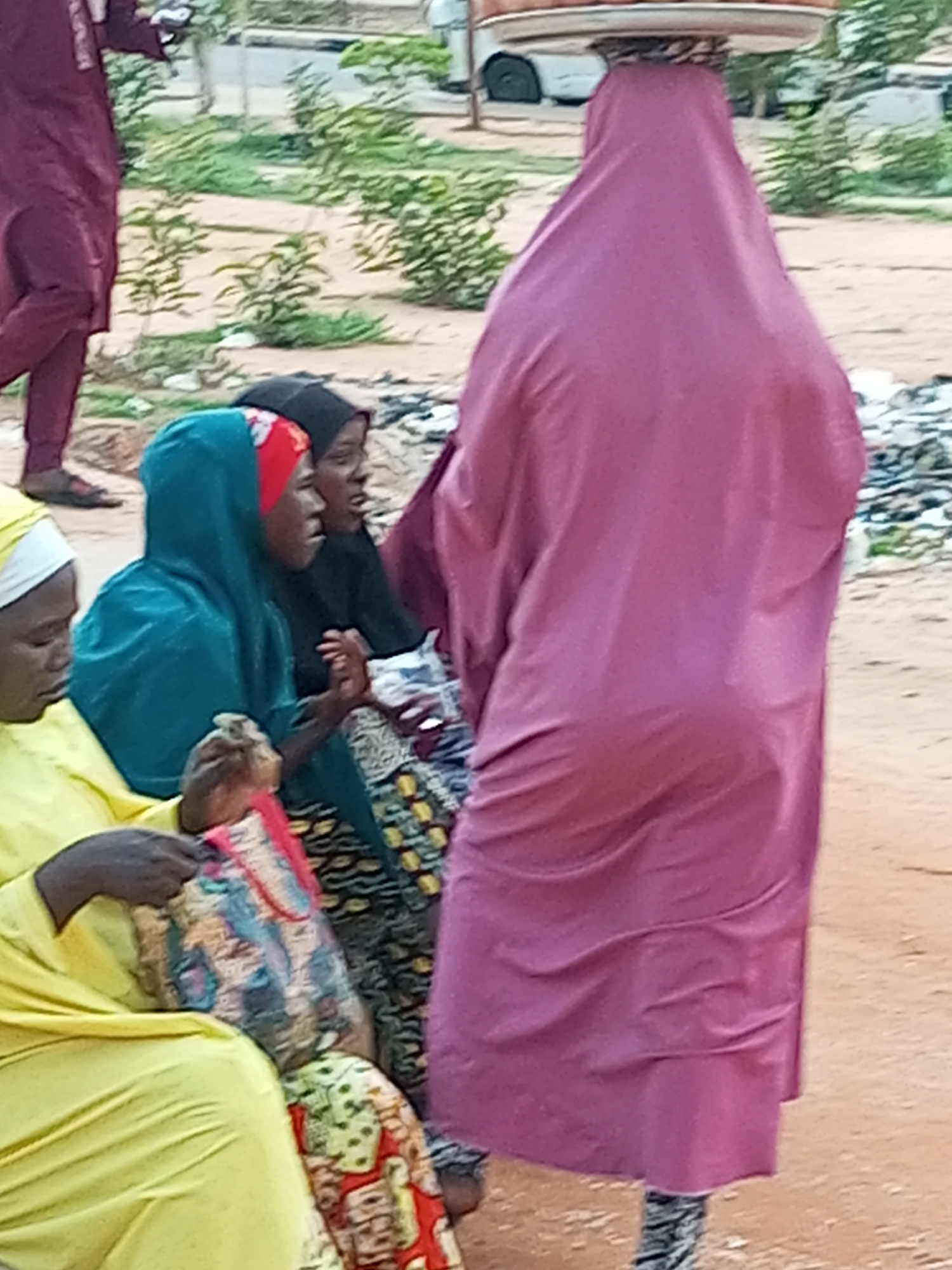 In Gombe, Nigeria, a group of individuals are gathered in an outdoor, dusty environment, possibly a market or roadside. A woman in a maroon garment stands on the left, facing away from the camera, with her arms held out. To her right, a person wearing a vibrant pink head covering and a flowing pink robe is the central focus, also facing away from the camera. This person is balancing a circular object on their head, which appears to be a tray or a makeshift plate.
In the foreground, three other individuals are visible, seated. On the far left, a person in a bright yellow garment and white head covering looks down at their hands. Next to them, to the right, is another person wearing a teal head covering and a red and white patterned scarf or inner headwear. This individual's hands are clasped together. Further to the right, partially obscured, is a person in a black head covering, looking to the side with an open mouth. They are holding what appears to be a small package or bag.
The ground is predominantly sand and dirt, with scattered pebbles and some dry vegetation. In the background, out-of-focus greenery and structures suggest an urban or semi-urban setting. The overall scene depicts a moment of everyday life, with individuals engaged in activities typical of a Nigerian community.