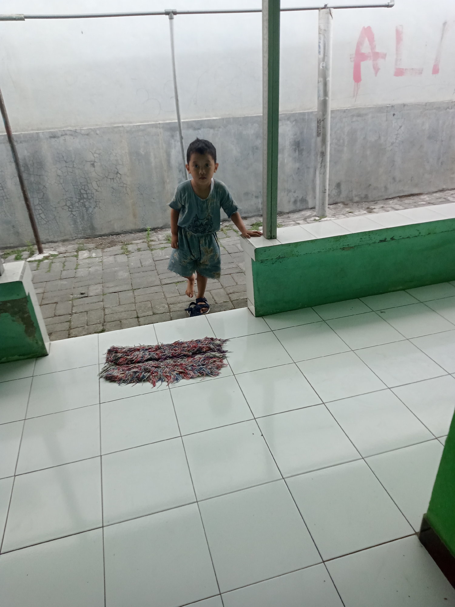 A young boy stands in an outdoor, covered walkway or patio area. The boy is wearing a light blue shirt and shorts and dark blue sandals. He is facing forward and looking at the camera. In front of him on the tiled floor is a small, colorful rug made of frayed threads. The walkway has a concrete wall in the background with some graffiti on it, and a green, raised concrete bench runs along one side. The floor is tiled with white square tiles. The area appears to be in Kedungturi, Indonesia, likely a residential or public space.