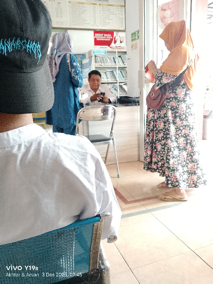 A person wearing a black cap and white shirt sits in a blue chair in the foreground. Behind them, a man in a white shirt uses a cell phone while sitting at a table. A woman in a blue dress and headscarf stands beside him. To the right, a woman in a floral dress and orange headscarf holds an object in her hand. A bookshelf and signs are visible in the background. The date and time "3 Des 2025, 07.45" are printed in the lower-left corner.