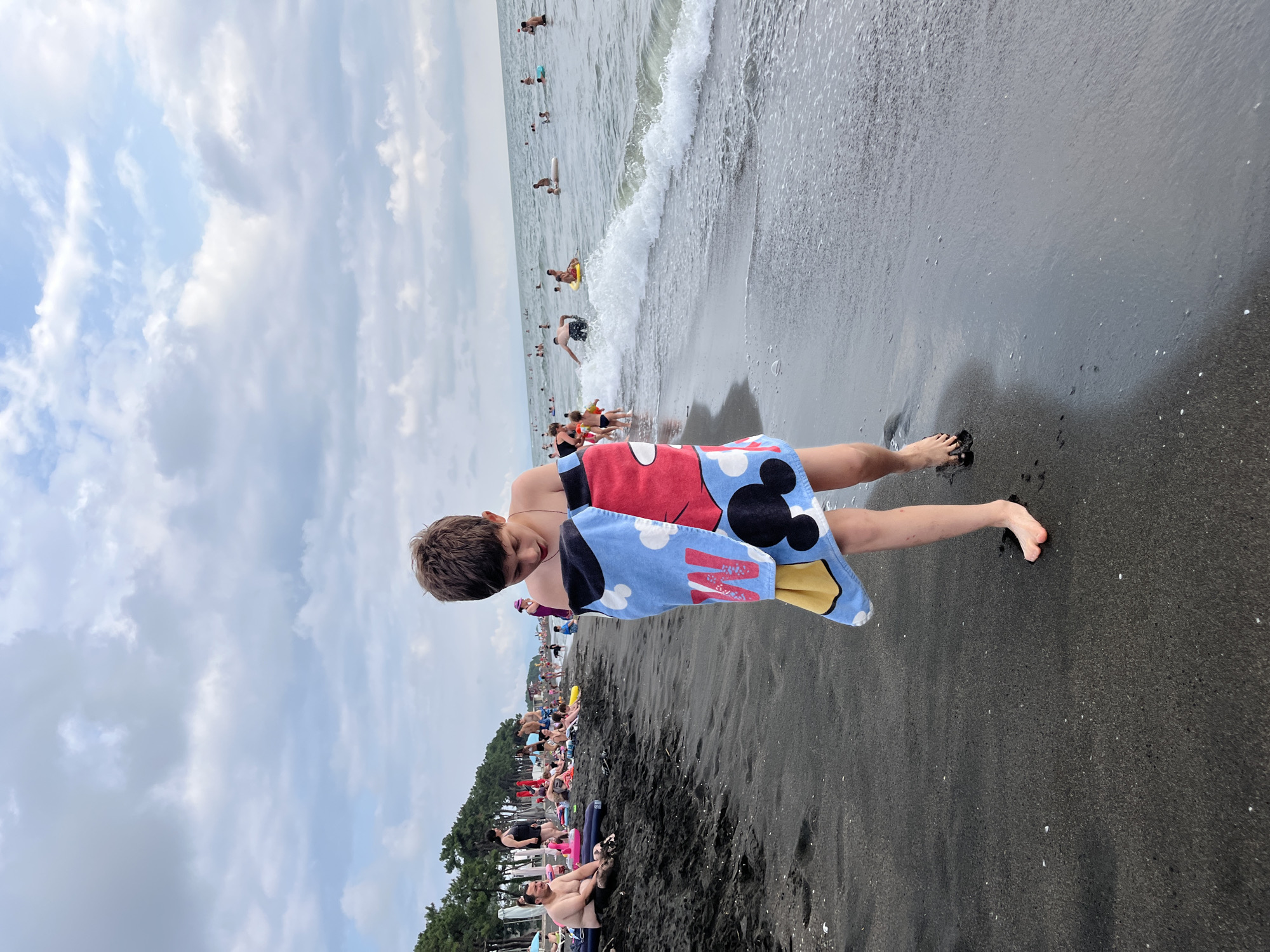 A young boy, wrapped in a towel with a Mickey Mouse design, walks on a black sand beach in Ureki, Georgia. He is looking down at his feet as he walks near the water's edge. In the background, other beachgoers are seen swimming and relaxing in the sea and on the shore. The sky is cloudy.