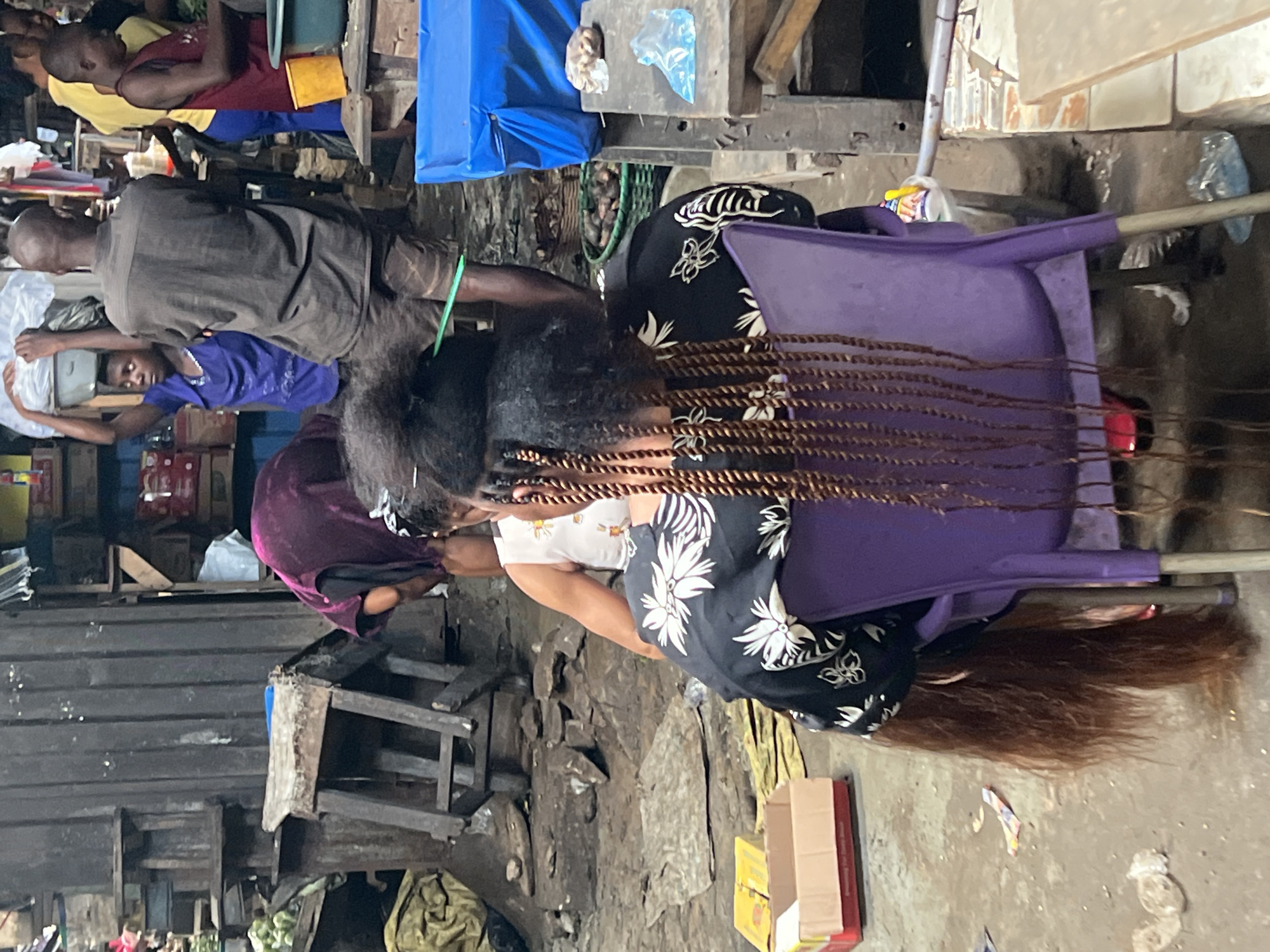 The image depicts an outdoor scene, likely a market or a workshop in Nkpor, Nigeria. Several individuals are present, engaged in various activities. In the foreground, a person's head and shoulders are visible, with their hair being braided. The braids are long and appear to be made of extensions. Behind this person, another individual is seated, wearing a black garment with a white floral pattern.
Further back, a group of people are seen in a more casual setting. One person wears a yellow shirt, another a maroon tank top over blue trousers, and a third person is wearing a blue t-shirt. They appear to be standing or sitting in a cluttered area with wooden structures and various objects, including what looks like blue tarpaulin and yellow containers.
The environment is somewhat disheveled, with visible debris and wooden planks scattered around. The overall impression is of a busy, informal setting. The lighting suggests it is daytime. There is no clear indication of the time of day beyond daylight. The weather is not discernible from the image.
Notable details include the intricate braiding of the hair in the foreground, the patterned clothing of the person seated, and the rustic nature of the surroundings with wooden elements and scattered items. The activity of hair braiding suggests a service being provided or a personal grooming session. The presence of multiple people in the background hints at a community or marketplace setting where such services might be offered or where people gather.
No visible text is clearly legible in the image. The scene is characteristic of a bustling environment in a Nigerian town, possibly a market stall or a workshop area. The emotions of the individuals are not clearly discernible due to the framing and focus of the image.