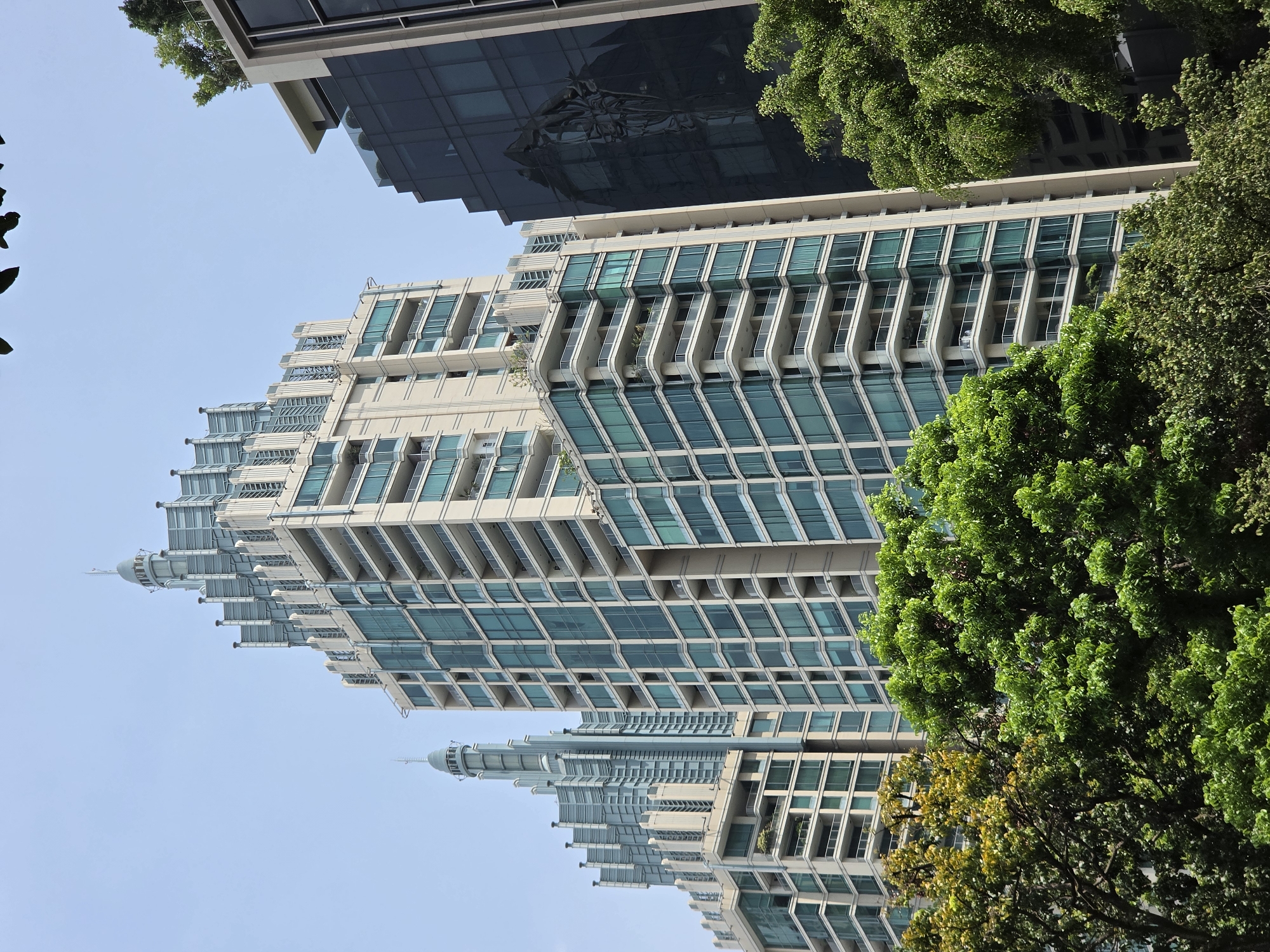 A low-angle shot of a tall, tiered building with many windows and balconies, taken in Bangkok, Thailand. The building is surrounded by lush green trees, some of which partially obscure the view. A dark glass building is partially visible in the upper left corner. The sky is a light blue color.
