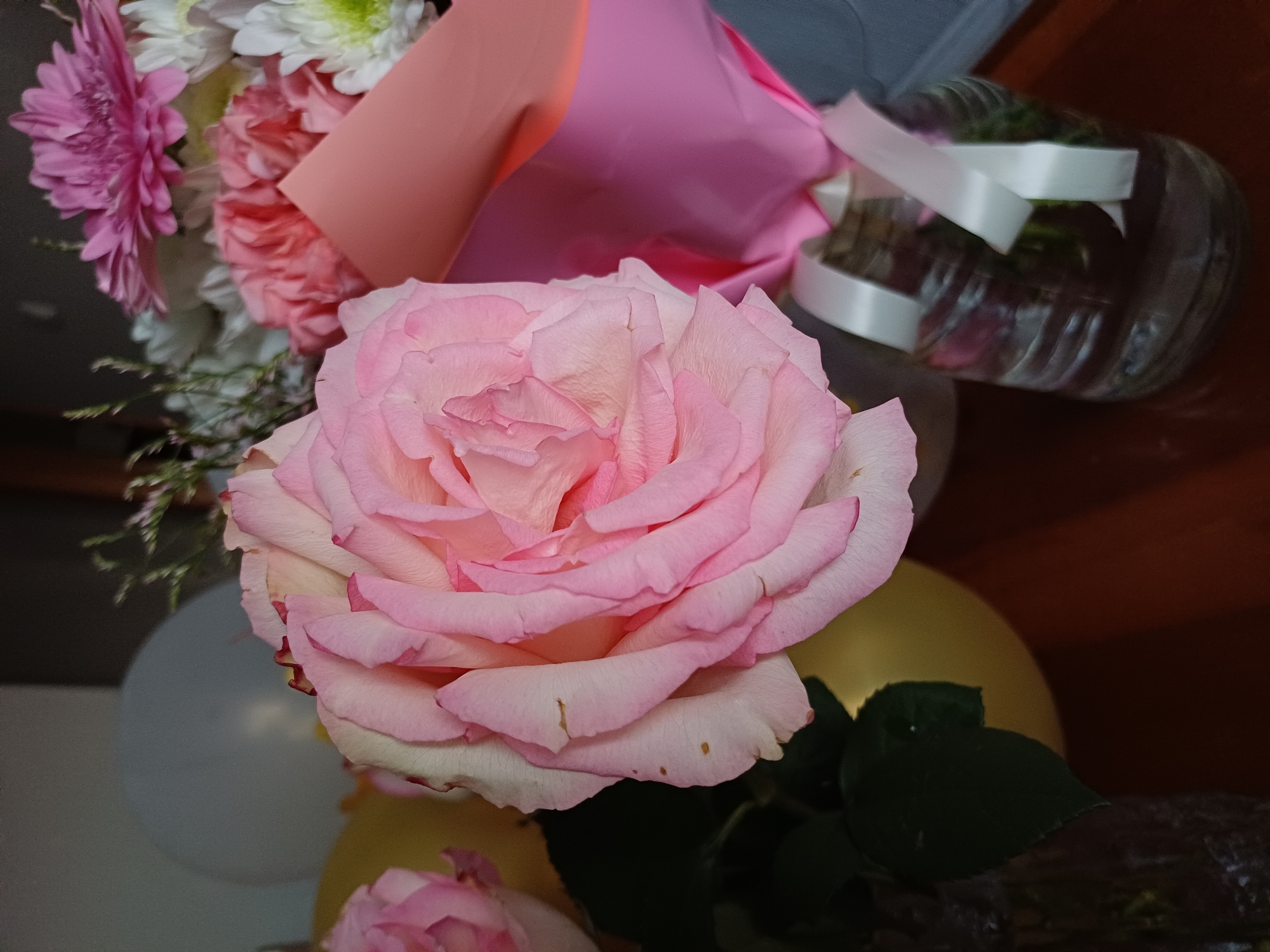 The image is a close-up of a bouquet of flowers, with a prominent pink rose in the foreground. The rose has soft, layered petals and a delicate gradient of color, ranging from a pale blush to a deeper pink at the edges. Behind the rose, several other flowers are visible, including pink carnations, white chrysanthemums, and a pink gerbera daisy. The flowers are wrapped in colorful paper, with shades of pink and peach.
In the background, there are several balloons, one a pale white and another a muted yellow. To the right, a clear plastic container with a white ribbon tied around it is visible, possibly a vase or a decorative element. The lighting suggests an indoor setting, with a warm, ambient glow, and the overall mood is celebratory or decorative. There is no visible text in the image. The scene appears to be a static arrangement of flowers, likely for an event or as a gift.