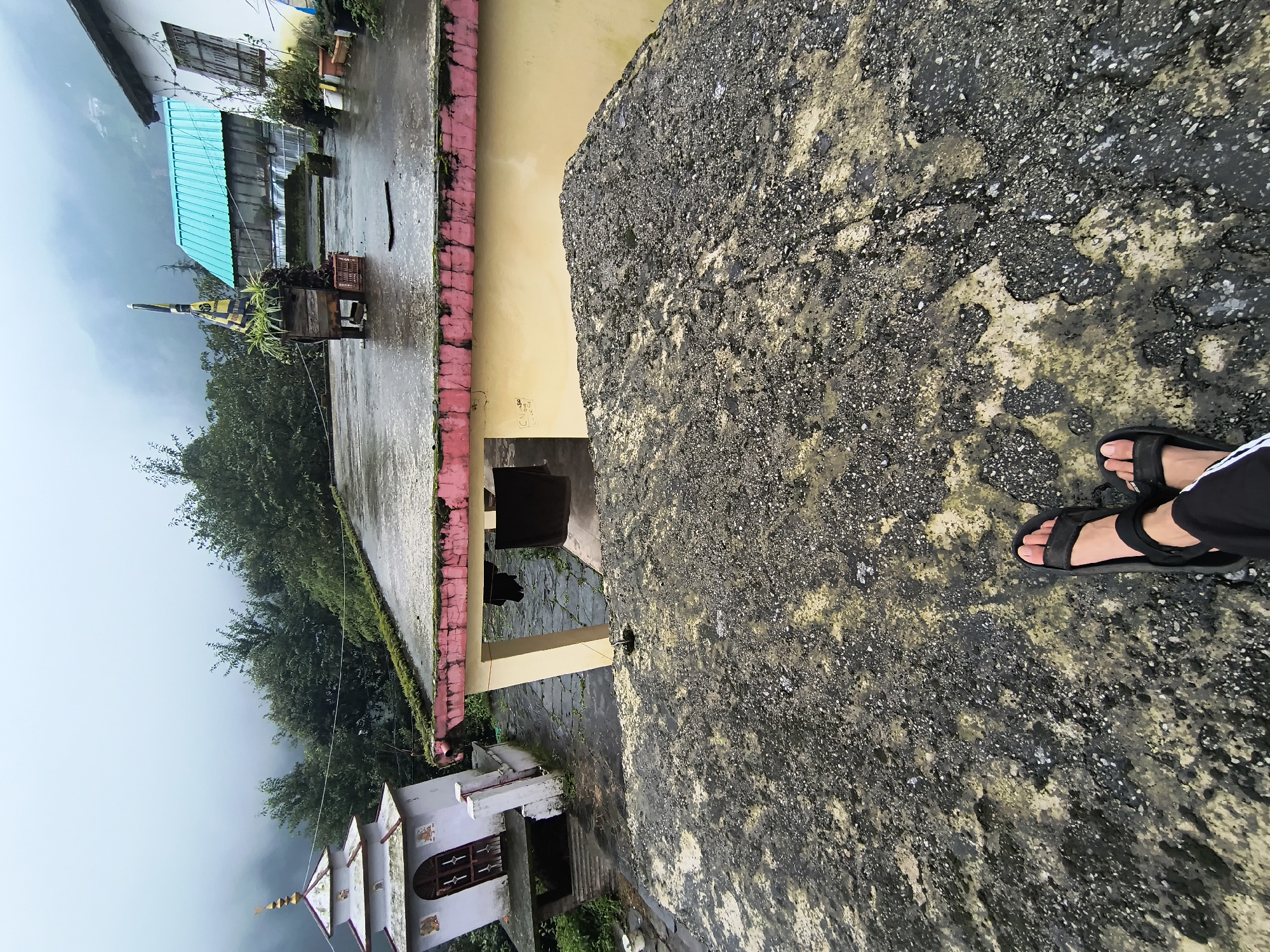 A person's feet wearing black sandals are visible in the foreground, standing on a rough, textured surface. The scene appears to be outdoors, possibly on a rooftop or elevated platform, overlooking a town or village nestled in a hilly, misty environment. A small, white temple with a spire is visible to the left, and a building with a teal-colored roof and white walls is situated in the middle ground. Trees and lush greenery are scattered throughout the landscape, and the sky is overcast with thick mist obscuring the distant mountains. The overall atmosphere is serene and somewhat isolated, suggesting a location in a rural or mountainous area.