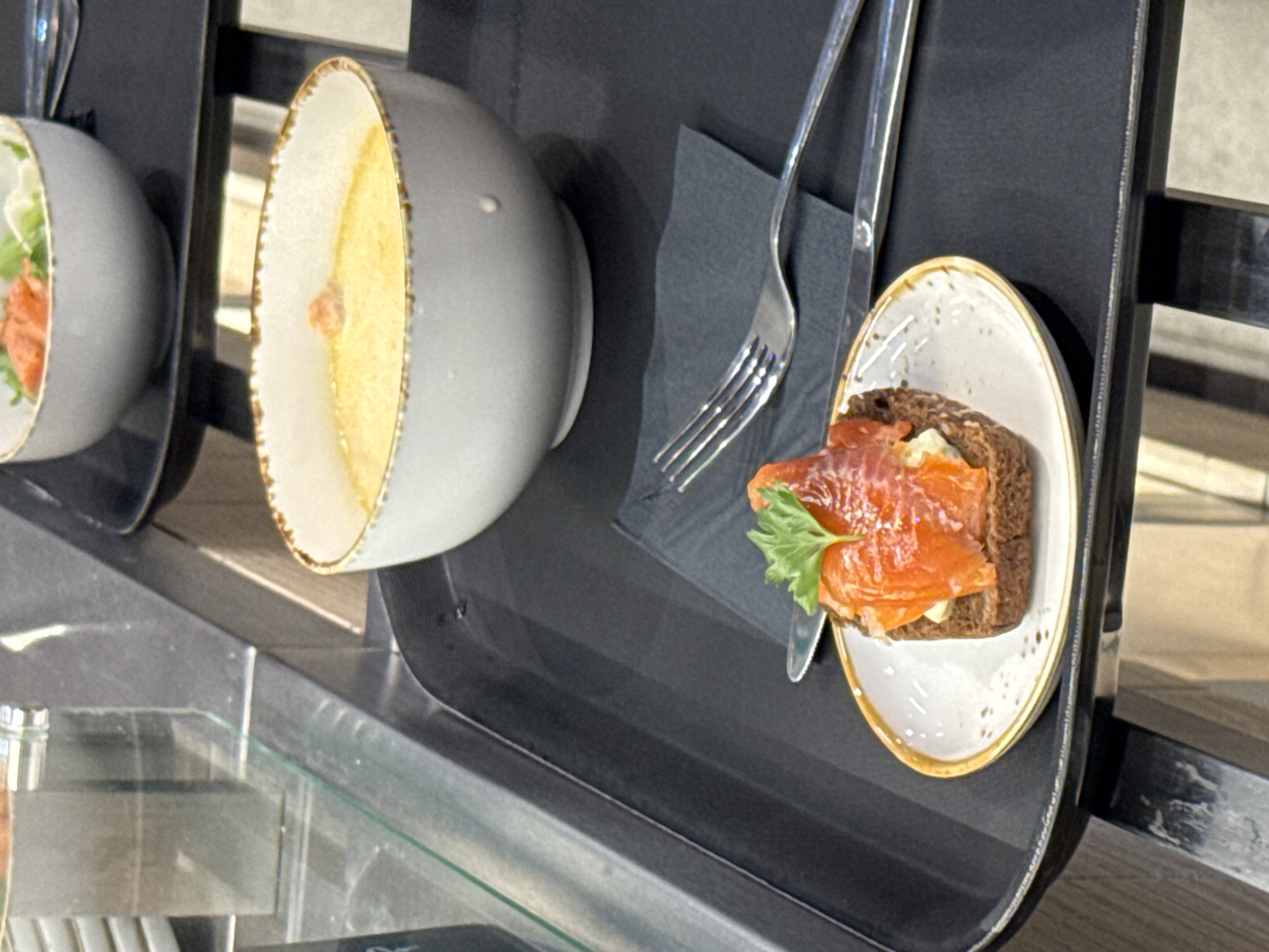 A close-up shot captures two bowls of soup on a dark tray. In the foreground, a small appetizer sits on a white plate with a gold rim. The appetizer is a slice of dark bread topped with what appears to be cream cheese and thinly sliced smoked salmon, garnished with a sprig of parsley. Next to the appetizer, a fork and knife are neatly placed on a black napkin. The soup in the foreground bowl is a creamy yellow color. In the background, another identical bowl of soup is visible, this one containing salmon and greens. The scene is set in a dimly lit environment with a reflective surface suggesting a display case or counter. The overall impression is one of a prepared meal, possibly in a restaurant or buffet setting. There is no specific information visible in the image to indicate a location in Yakkima, Russia.