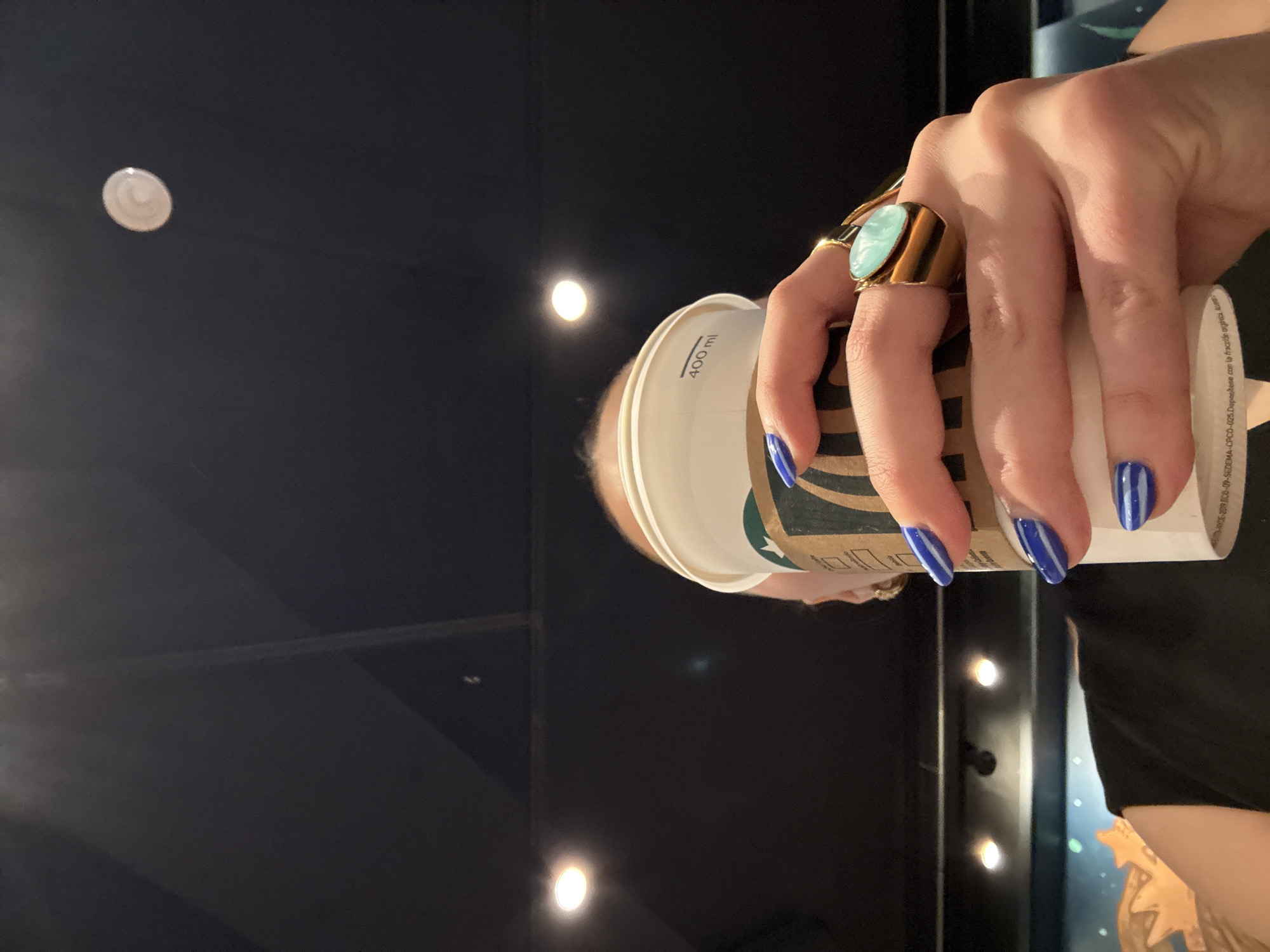 A close-up, low-angle shot depicts a person holding a white Starbucks paper cup with their right hand, obscuring most of their face. The hand displays blue fingernail polish with white horizontal stripes and a large gold-toned ring with an oval, light-blue or turquoise-colored stone on the ring finger. The cup features a brown Starbucks sleeve fragment and "400 ml" printed near the top rim. The bottom edge of the cup includes partially visible text in Spanish, "SEDEPA-CPCO-025. Deposítese con la fracción orgánica. Autoriz...", indicating the cup is intended for composting in Mexico. This detail places the scene within Mexico City, Mexico.
A small portion of the person's forehead and hairline is visible above the cup, along with the shoulder and upper arm of a dark-colored garment, possibly a black sleeveless top. A faint gold earring is visible near the person's right ear. The background is predominantly dark, with several bright, circular recessed ceiling lights. Below the lights, a dark teal-blue wall or surface with abstract, lighter-colored shapes, possibly depicting celestial bodies or stars, is visible on the lower left.