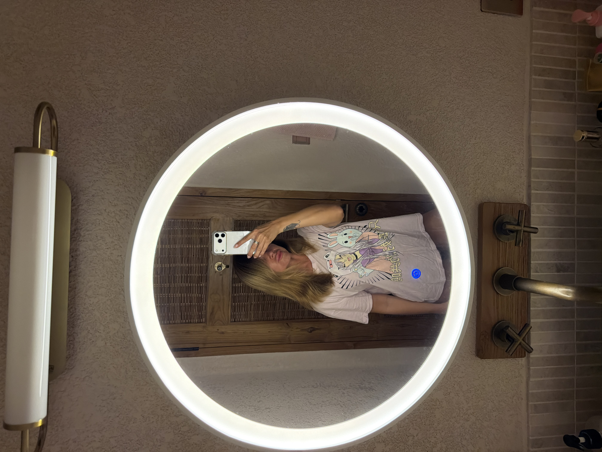 A woman takes a mirror selfie in a bathroom. She is wearing a light pink t-shirt with a graphic print and the reflection of her phone is visible in the mirror. The bathroom features a large circular mirror with a built-in light, a wooden vanity with faucets, and textured walls. The background shows a wooden door and a tiled backsplash. The scene appears to be in Mengwi, Indonesia.