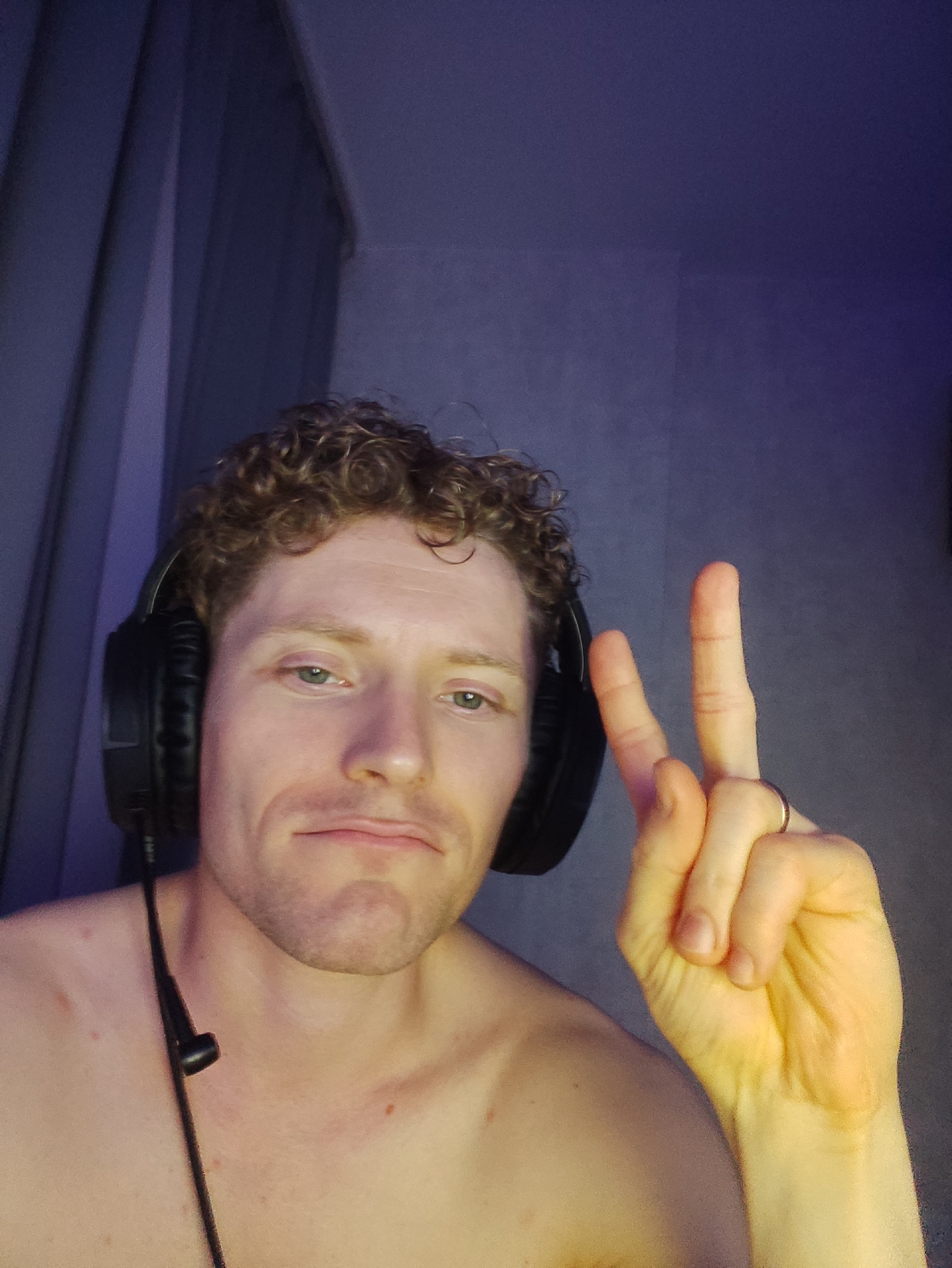 A pale-skinned man with curly blonde hair and green eyes is shown from the chest up. He is wearing black headphones with a microphone and making a peace sign with his right hand. He is shirtless and has a ring on his right ring finger. The background is dark and consists of purple curtains and a textured wall. The lighting is dim and appears to be artificial. The image quality is grainy.
