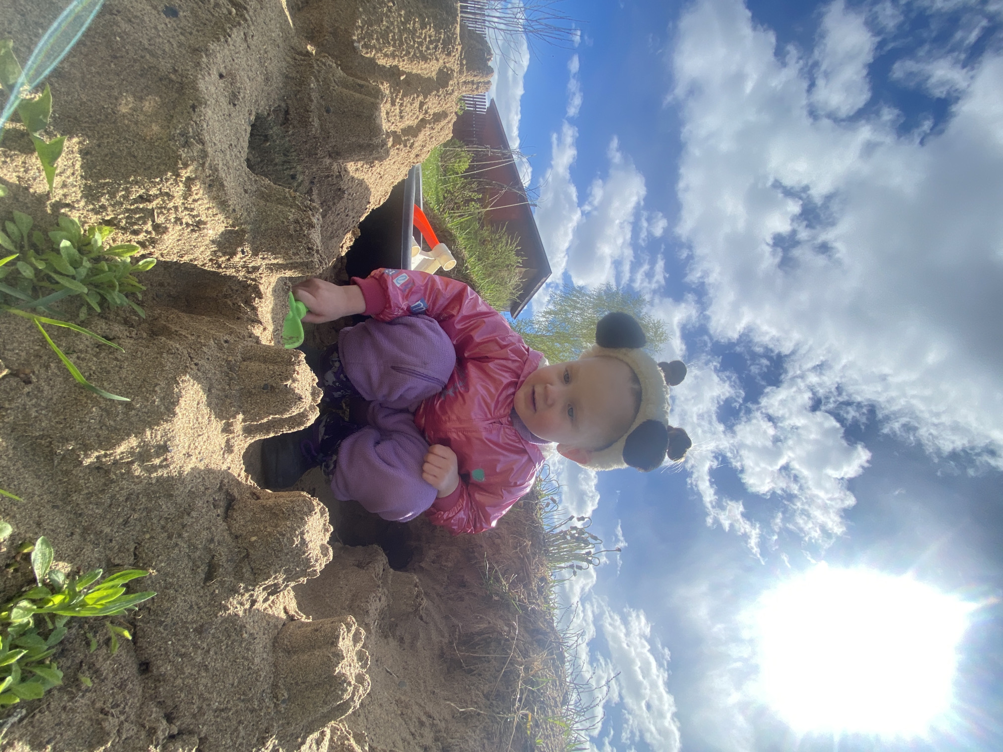 A young child wearing a pink jacket and purple pants is playing in a sandpit. The child also has a headband with ears. The sandpit appears to be fairly large, with tall mounds of sand. The child is holding a green plastic toy in their hand. The background includes a blue sky with clouds and the sun shining brightly. The location is not evident from this image.