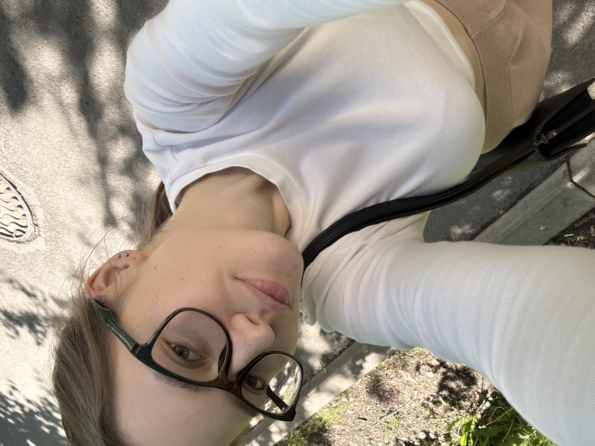The image is a tilted, eye-level selfie featuring a young person outdoors, reportedly in Saint Petersburg, Russia. The individual, likely female, has light skin, brown hair, and is wearing dark-framed glasses. Two helix piercings are visible on their left ear. They are dressed in a white long-sleeved top and a beige or brown lower garment, with a black strap crossing their chest, likely from a black clutch bag visible near their hip.
The person is looking directly into the camera with a neutral expression. The setting appears to be a paved area, possibly a sidewalk or street, with light-colored asphalt or concrete ground. Strong shadows of tree branches or leaves are cast across the ground, indicating a sunny daytime. Patches of dirt and green foliage are visible on the right side of the frame. A metal manhole cover with a distinctive wavy pattern is partially visible on the left. No discernible text is present in the image.