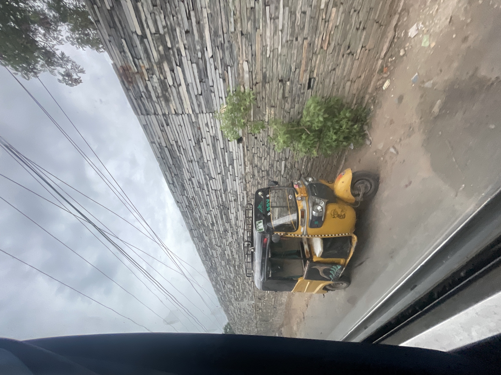 The image shows a yellow auto-rickshaw parked on a dirt road next to a stacked stone wall. The sky is overcast with visible power lines. A small green bush grows in front of the wall. The auto-rickshaw has a black roof and is partially open. The scene appears to be in Bethamcherla, India.